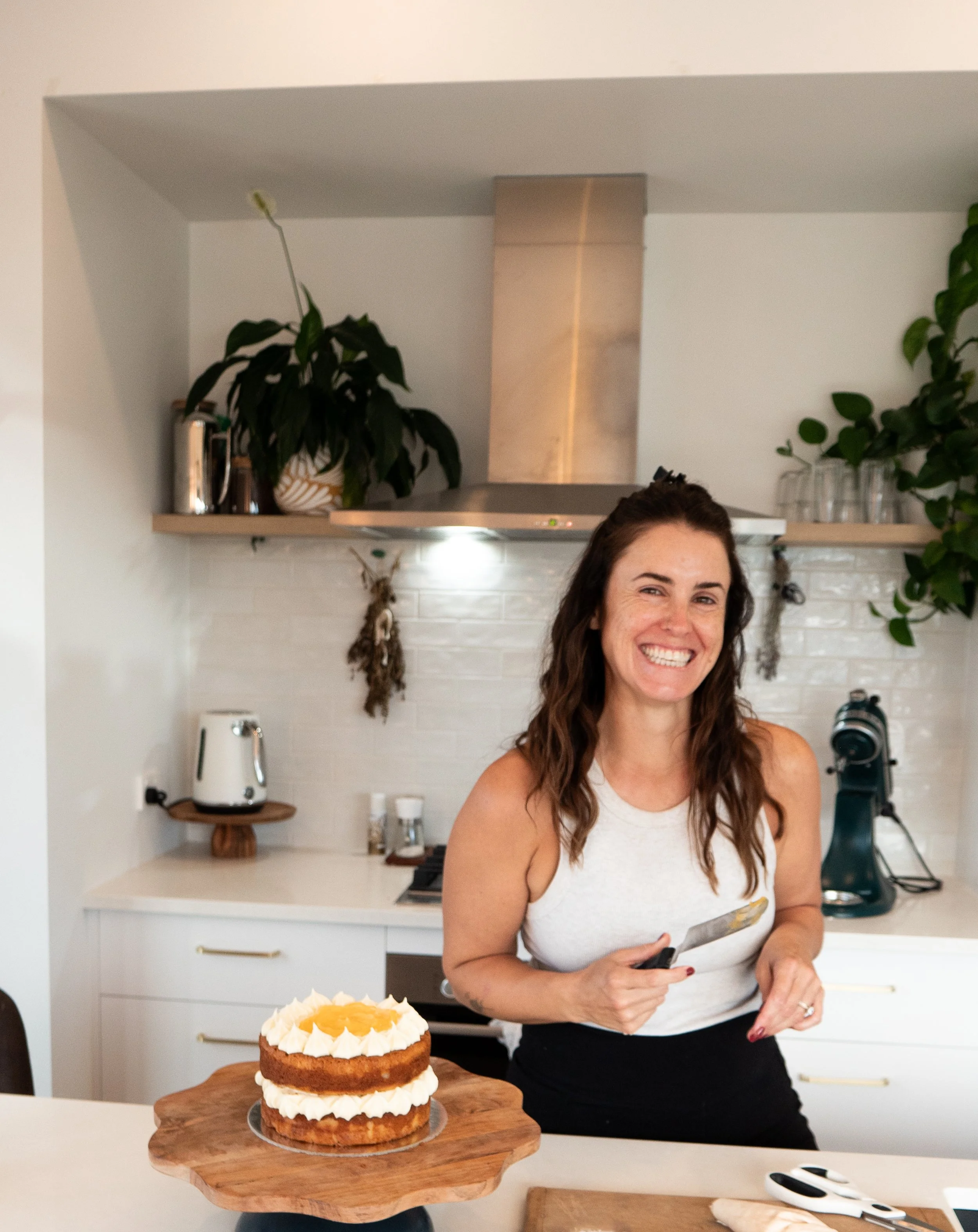 A woman with brown hair wearing a white tank top and black pants, smiling and holding a knife, standing in a kitchen near a cake on a wooden cake stand.
