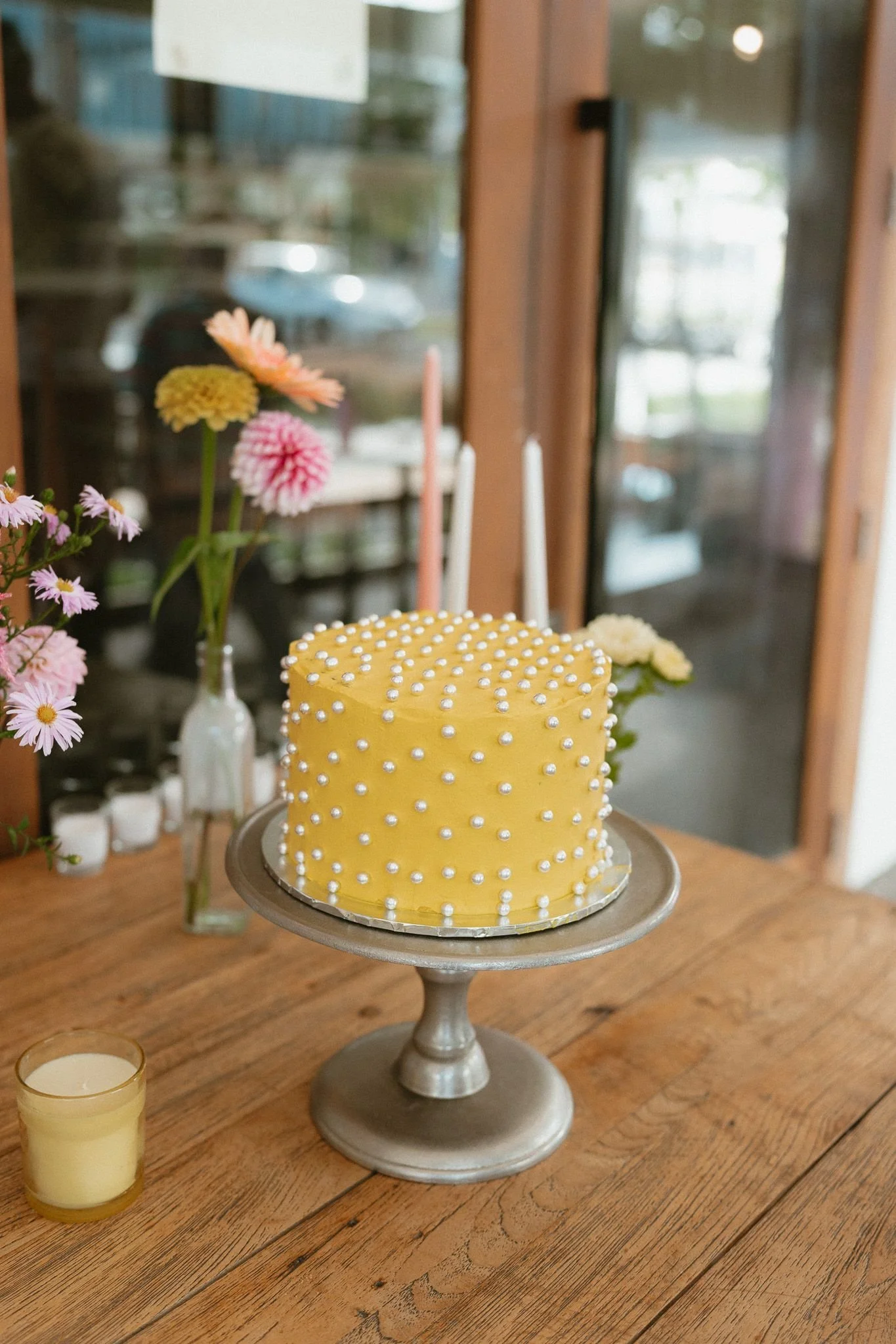 A yellow birthday cake decorated with white pearl-like sprinkles on a silver cake stand, placed on a wooden table with pink and white flowers and candles in the background.