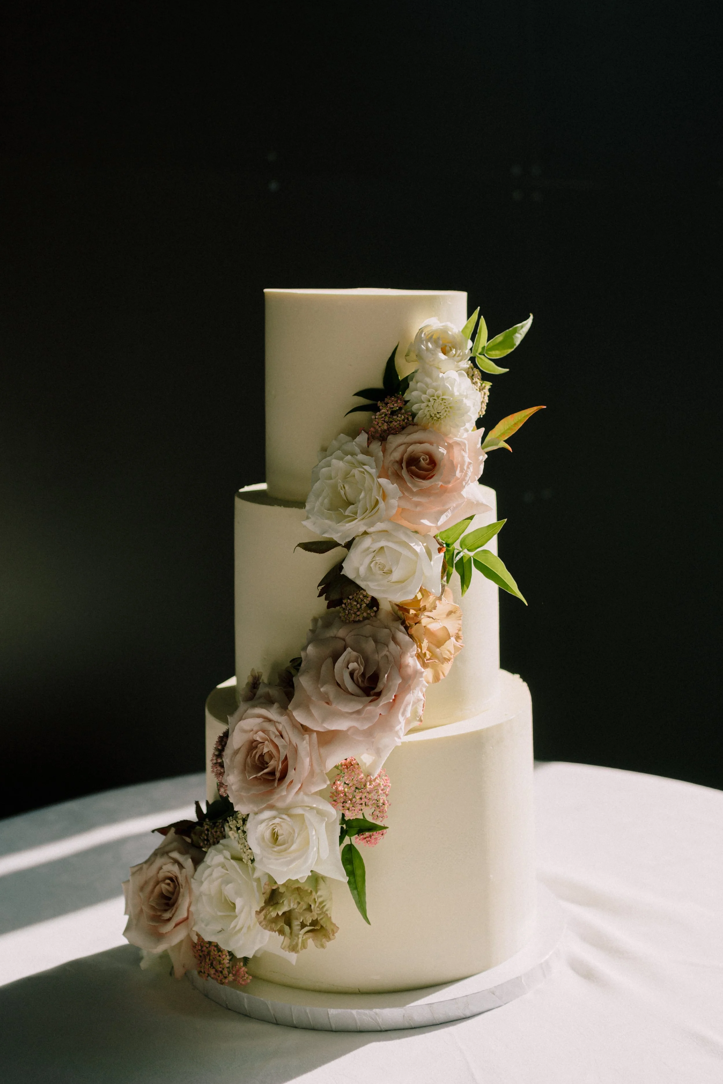 A three-tiered wedding cake decorated with cascading white, pink, and lavender roses and green leaves, placed on a white table.