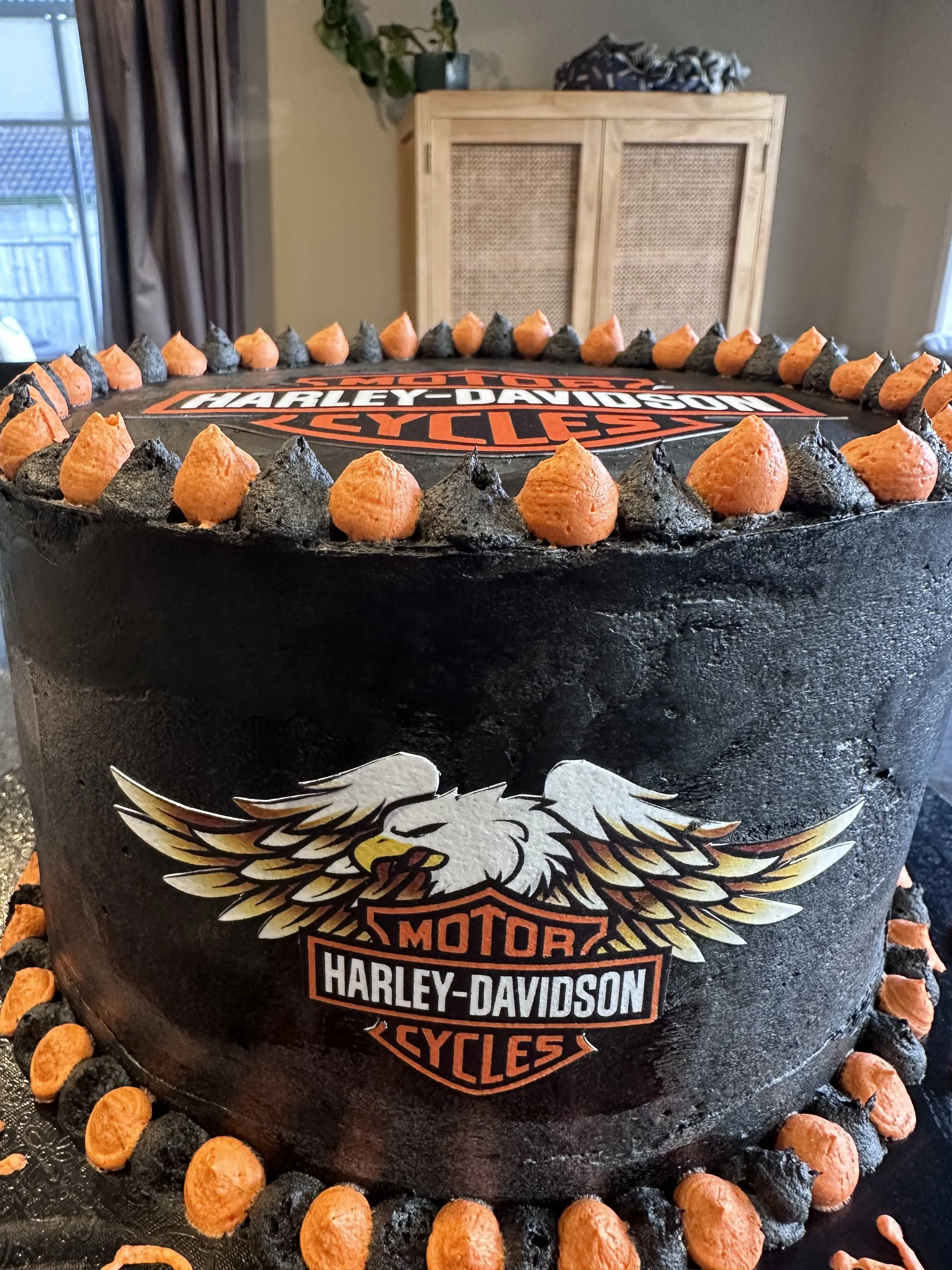 Harley-Davidson themed birthday cake with black icing and orange and black piped decorating tips around the top and bottom edges; features the Harley-Davidson logo with a bald eagle on the front.