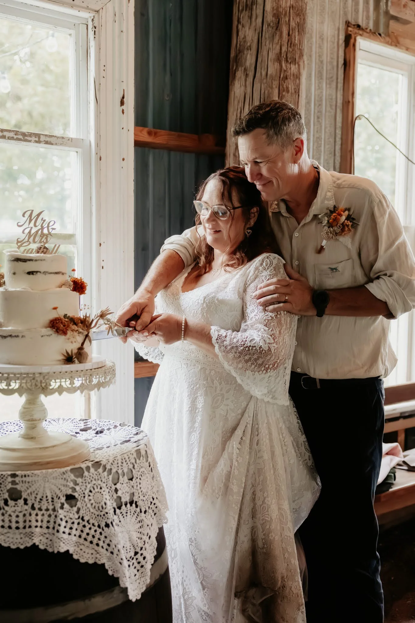 A woman in a lace wedding dress and a man in a light shirt are cutting a wedding cake together, standing in a rustic room with large windows.