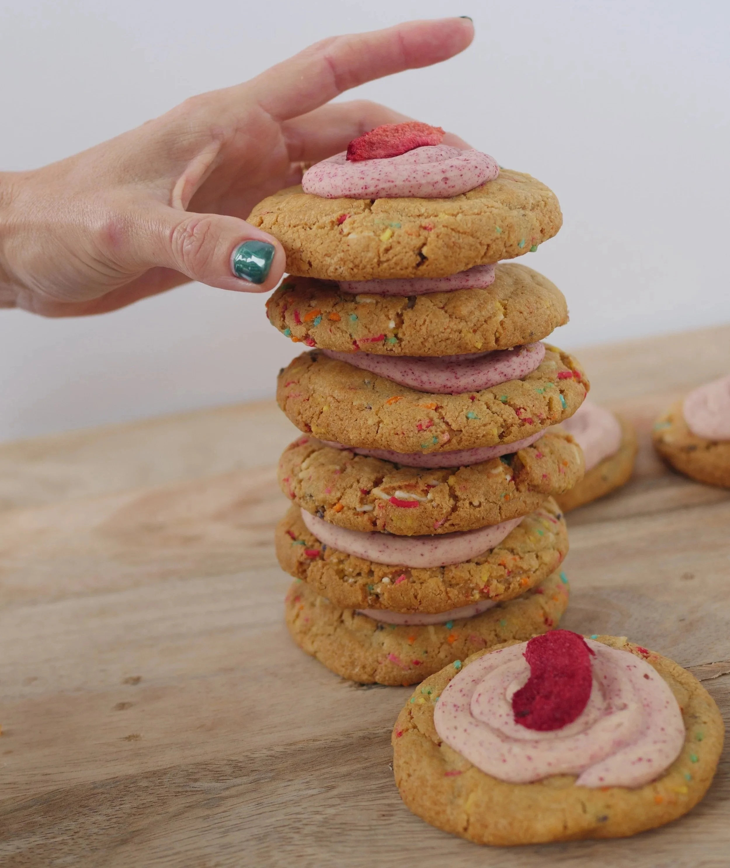 A hand holding a stack of six large, colorful cookies with pink frosting in the middle and topped with a small piece of candy. There are two more cookies with pink frosting and candy on a wooden surface nearby.