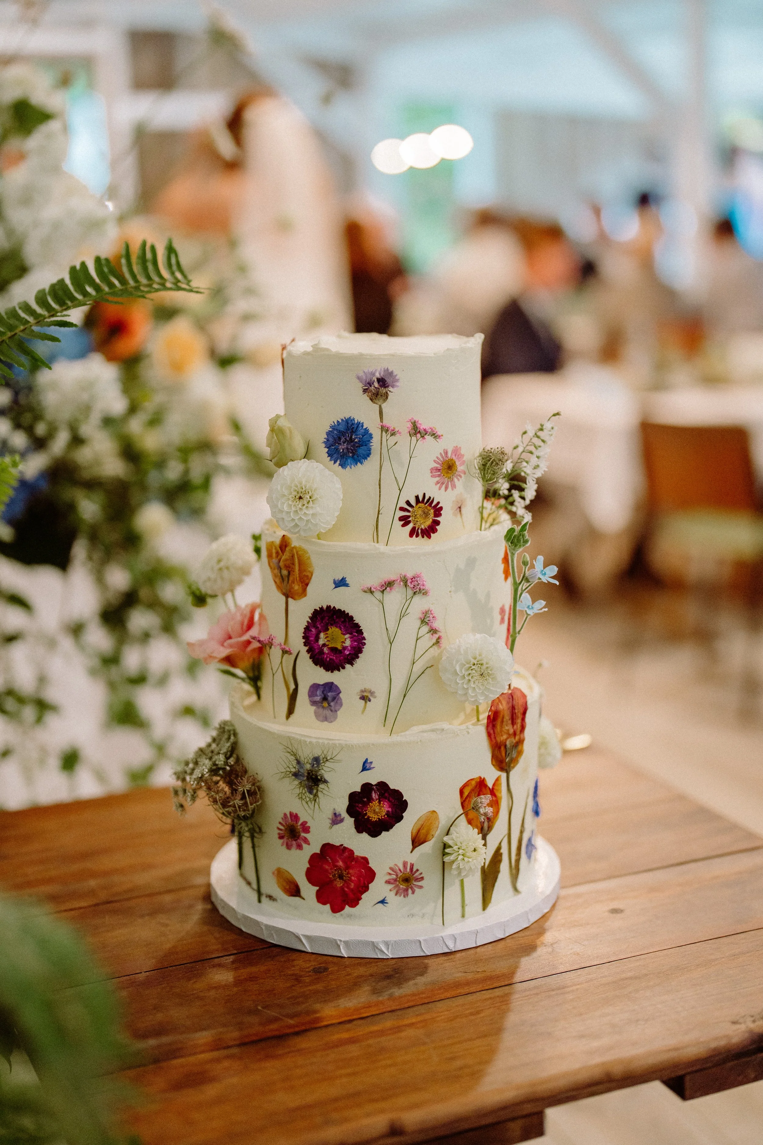 Three-tiered wedding cake decorated with pressed and dried flowers on a wooden table, with blurred wedding reception in the background.