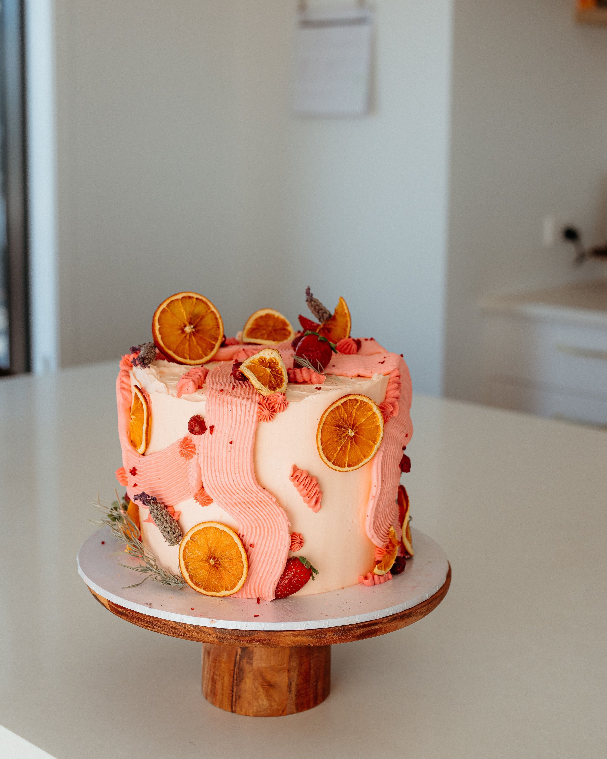 A decorated cake topped with dried orange slices, strawberries, and pink frosting, set on a white and wood cake stand on a white table.