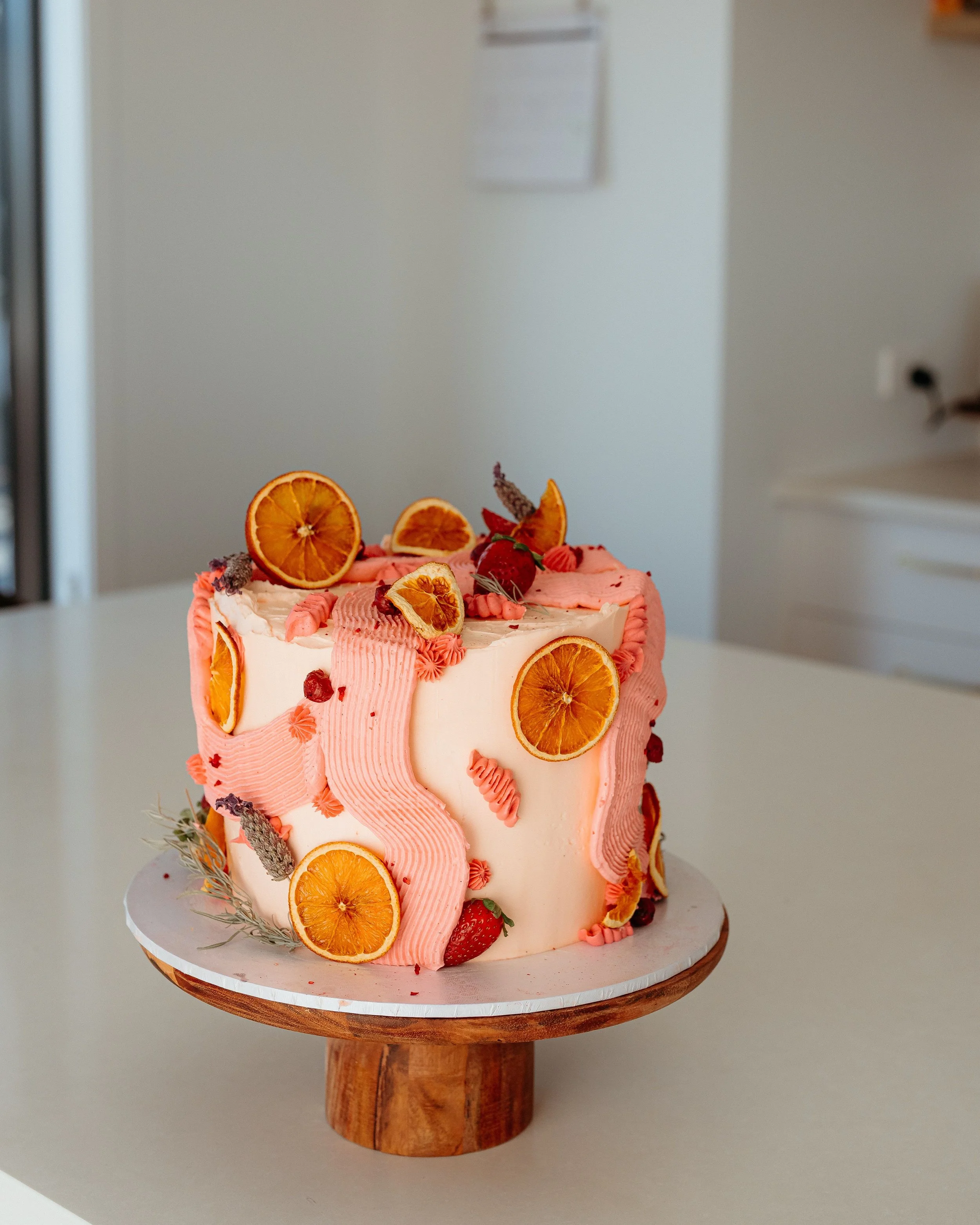 A pink and white decorated cake with dried orange slices and strawberries on top, placed on a wooden cake stand.