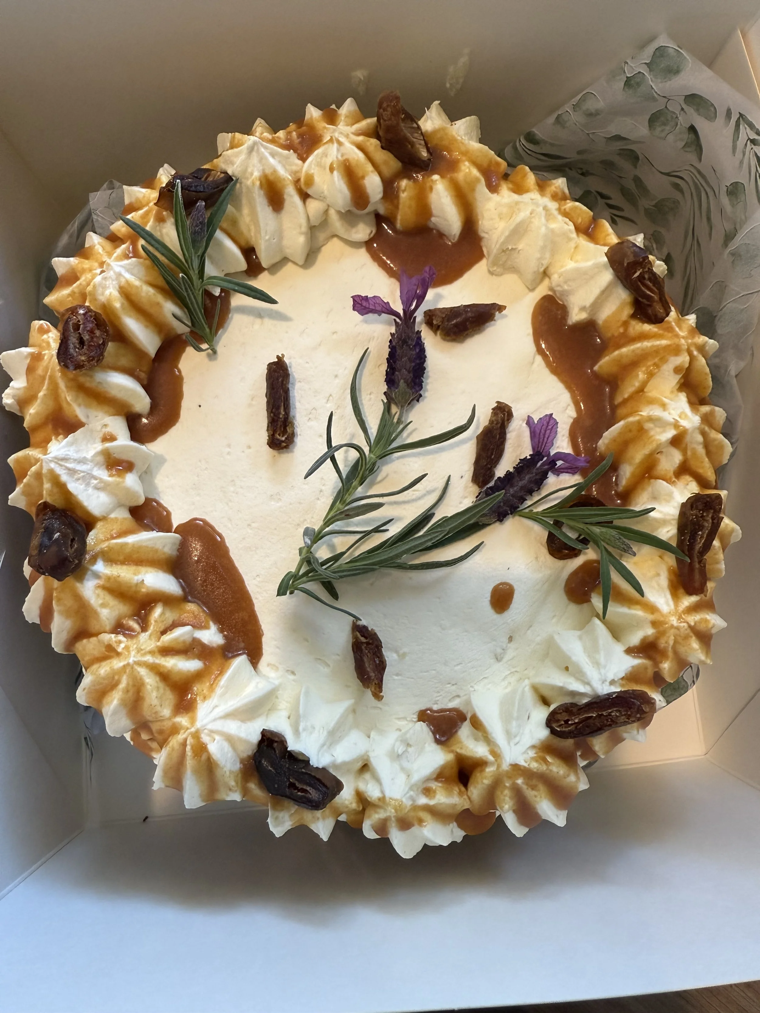 Round cake with white icing, decorated with caramel drizzle, whipped cream rosettes, dates, and sprigs of rosemary and lavender.