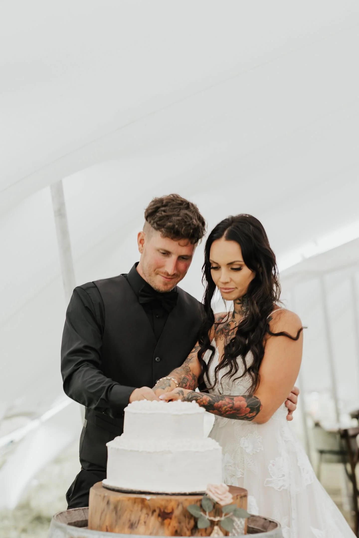 A newlywed couple cutting a wedding cake under a white tent with the bride's tattoos visible.
