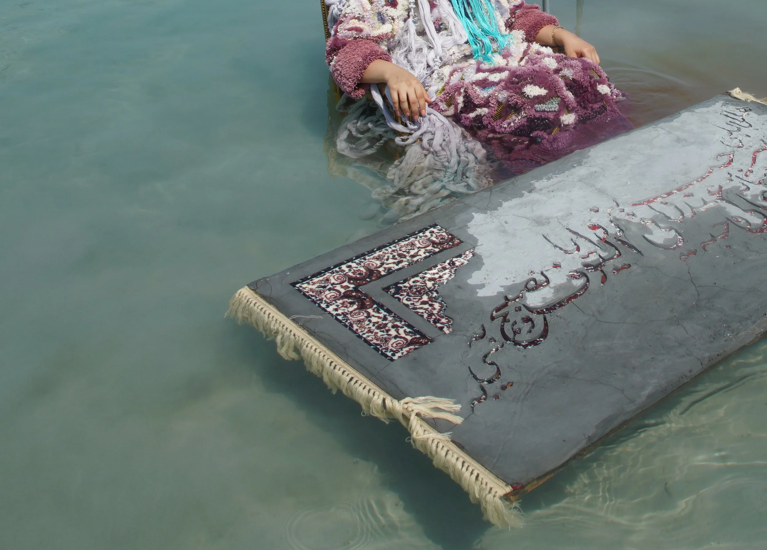 A person dressed in colorful clothing sitting on a submerged altar in water, with part of the altar visible above the water surface, featuring a patterned cloth and inscriptions. Ayesha Hadhir, Rawdha Al Ketbi and Shaikha Al Ketbi.