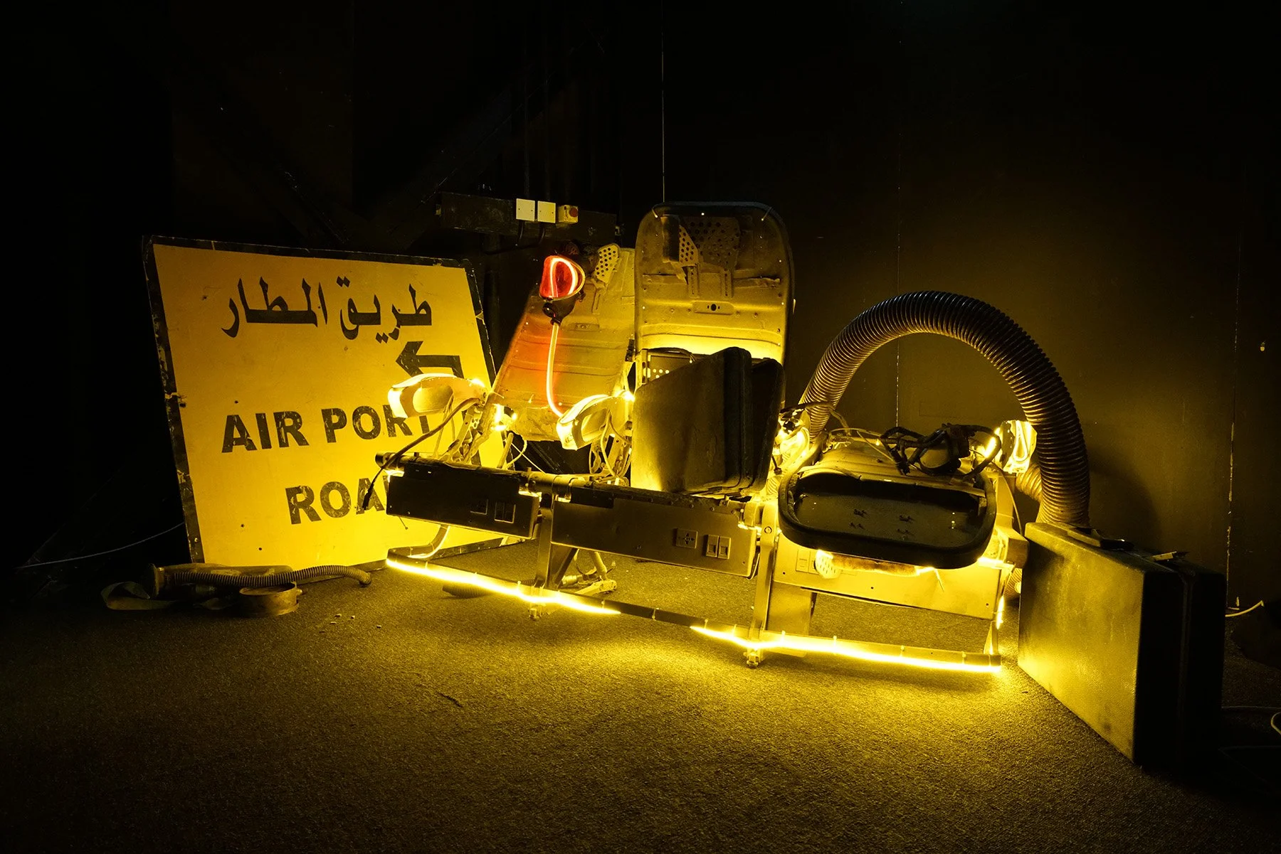 A broken airport runway sign with Arabic and English text, a damaged black airplane seat, illuminated by yellow lighting, with various cables and a hose in a dark background.