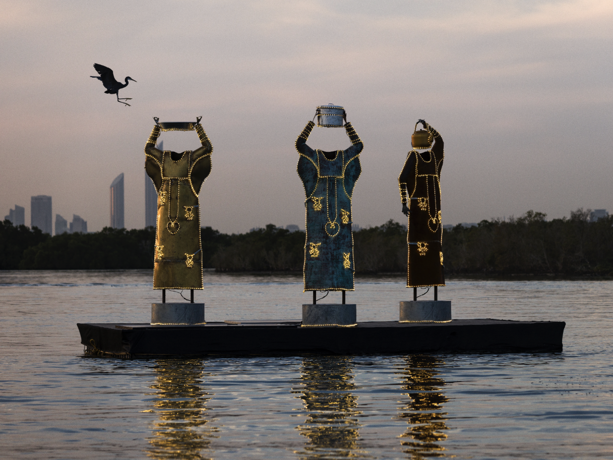Art installation of three dress sculptures with illuminated outlines, floating on water at sunset, with city skyline and a bird flying overhead. Ayesha Hadhir, Rawdha Al Ketbi and Shaikha Al Ketbi.