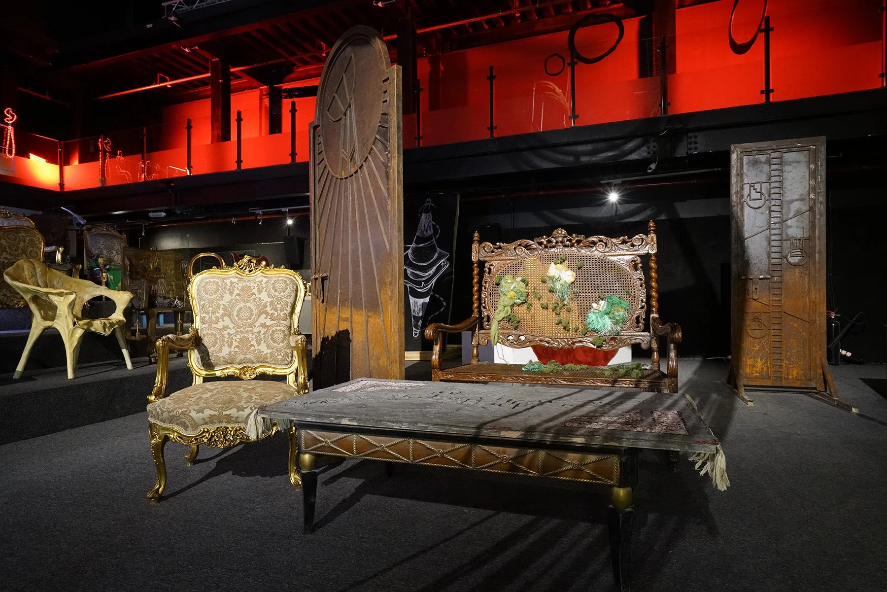 A room with vintage and modern furniture including an ornate gold throne-like chair, an intricate wooden bench, a gilded armchair, and sculptural chairs, set against a dark background with red lighting and decorative items.