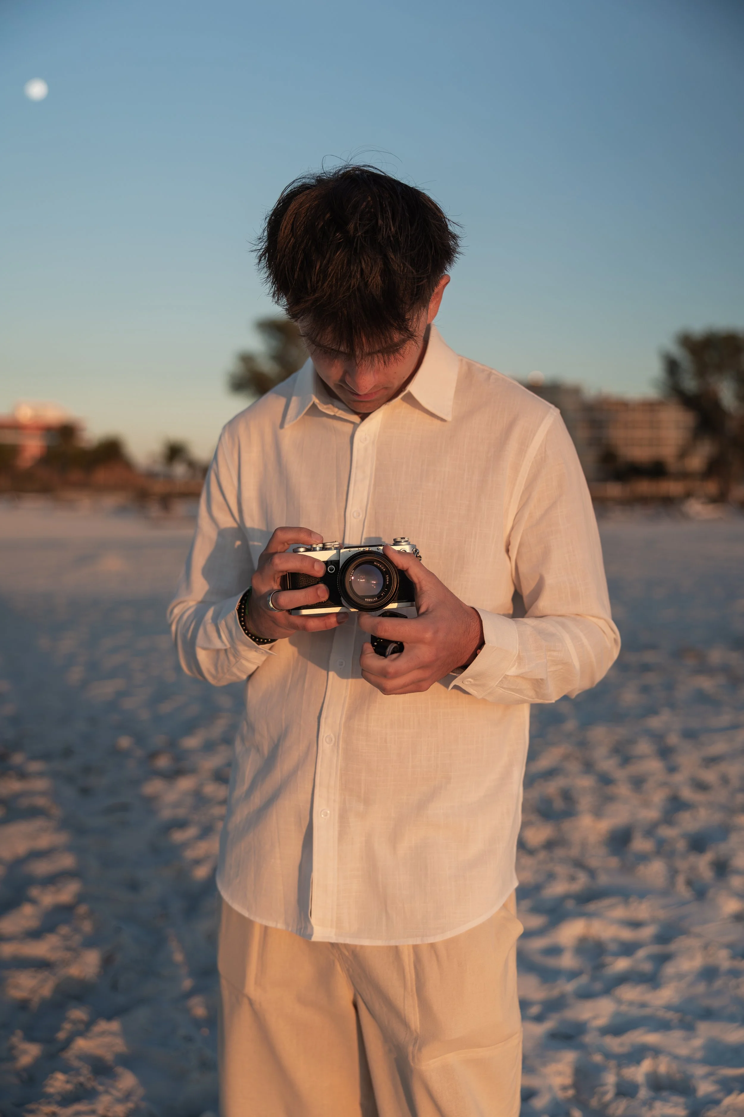 A young man on a beach holding a camera, looking down at it, during sunset or golden hour.