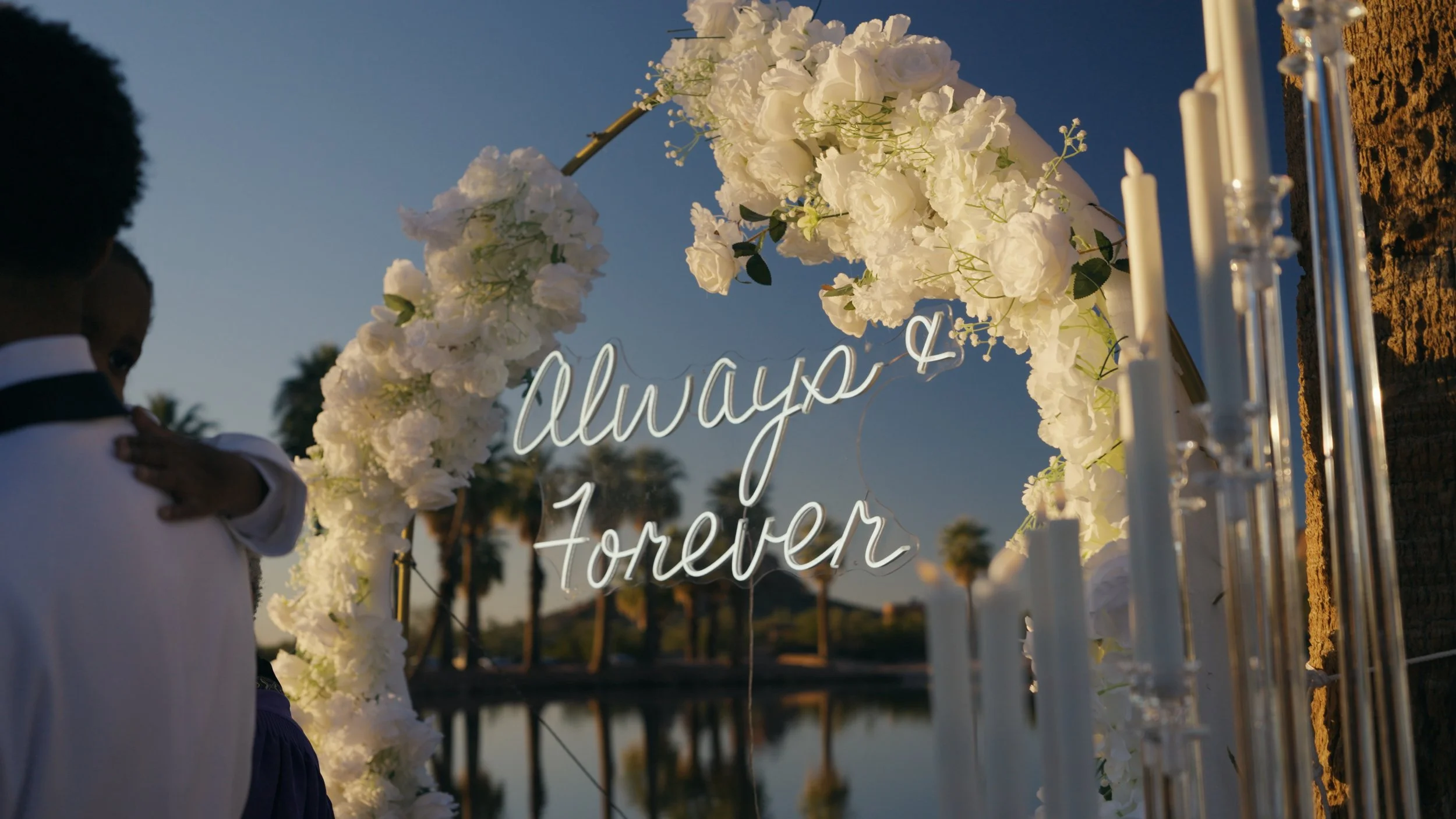 A wedding decoration with white flowers and a neon sign that reads 'Always & Forever' near a body of water with trees in the background during sunset.