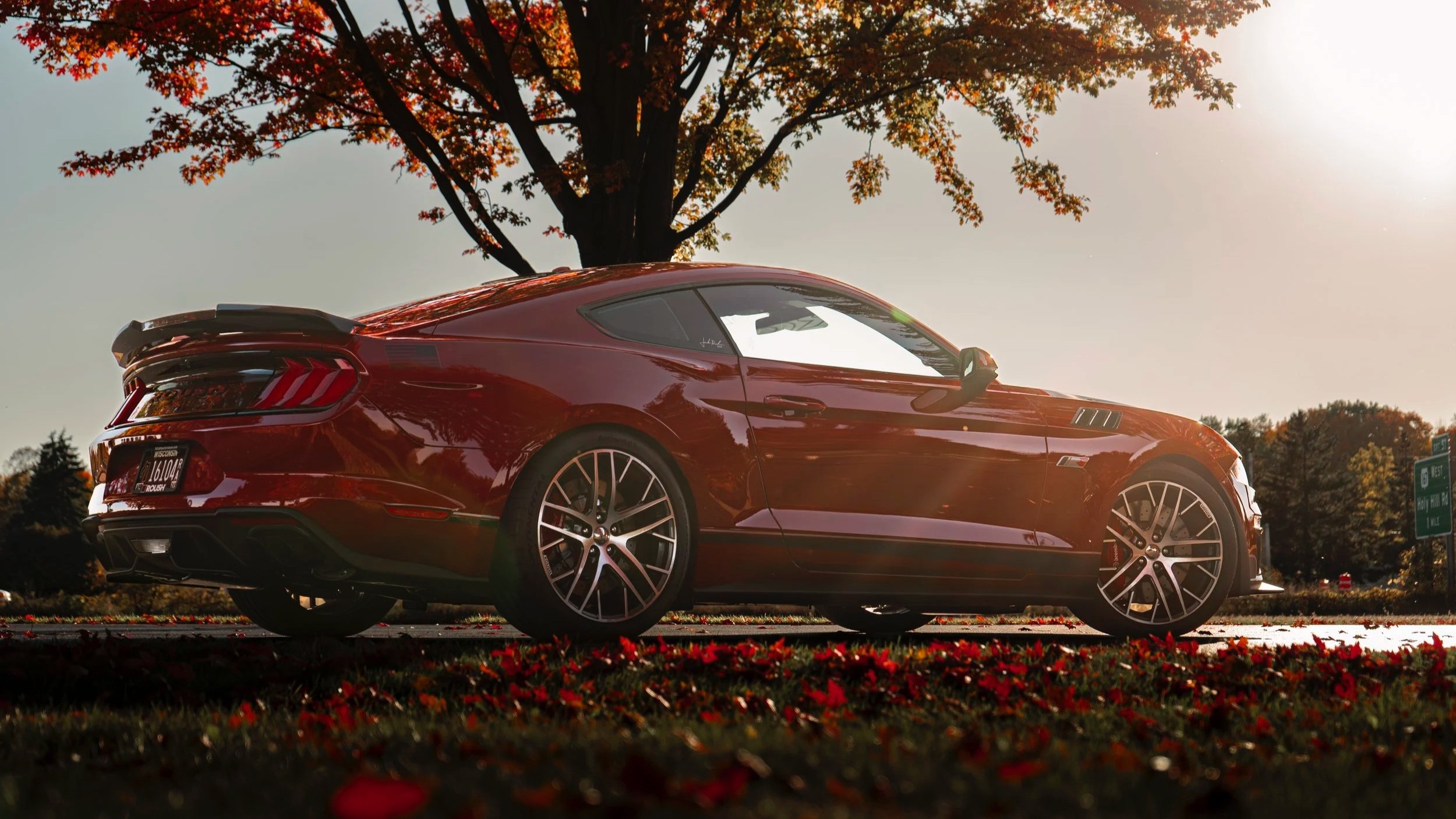 Red sports car parked on a road with fallen autumn leaves and trees in the background during sunset.