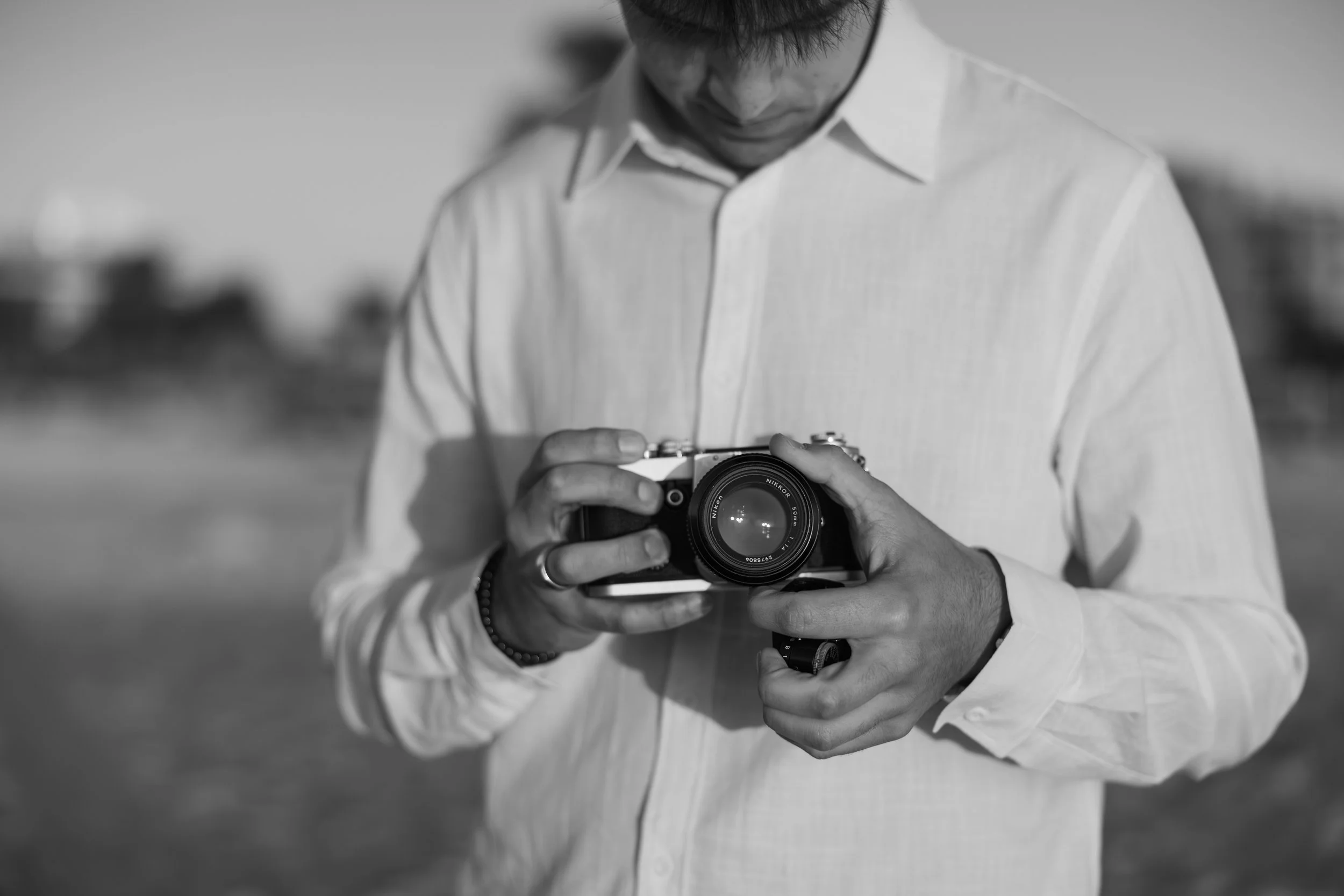 A person in a light-colored shirt holds a camera in both hands, looking down at it. The background is blurred, suggesting an outdoor setting, and the image is in black and white.