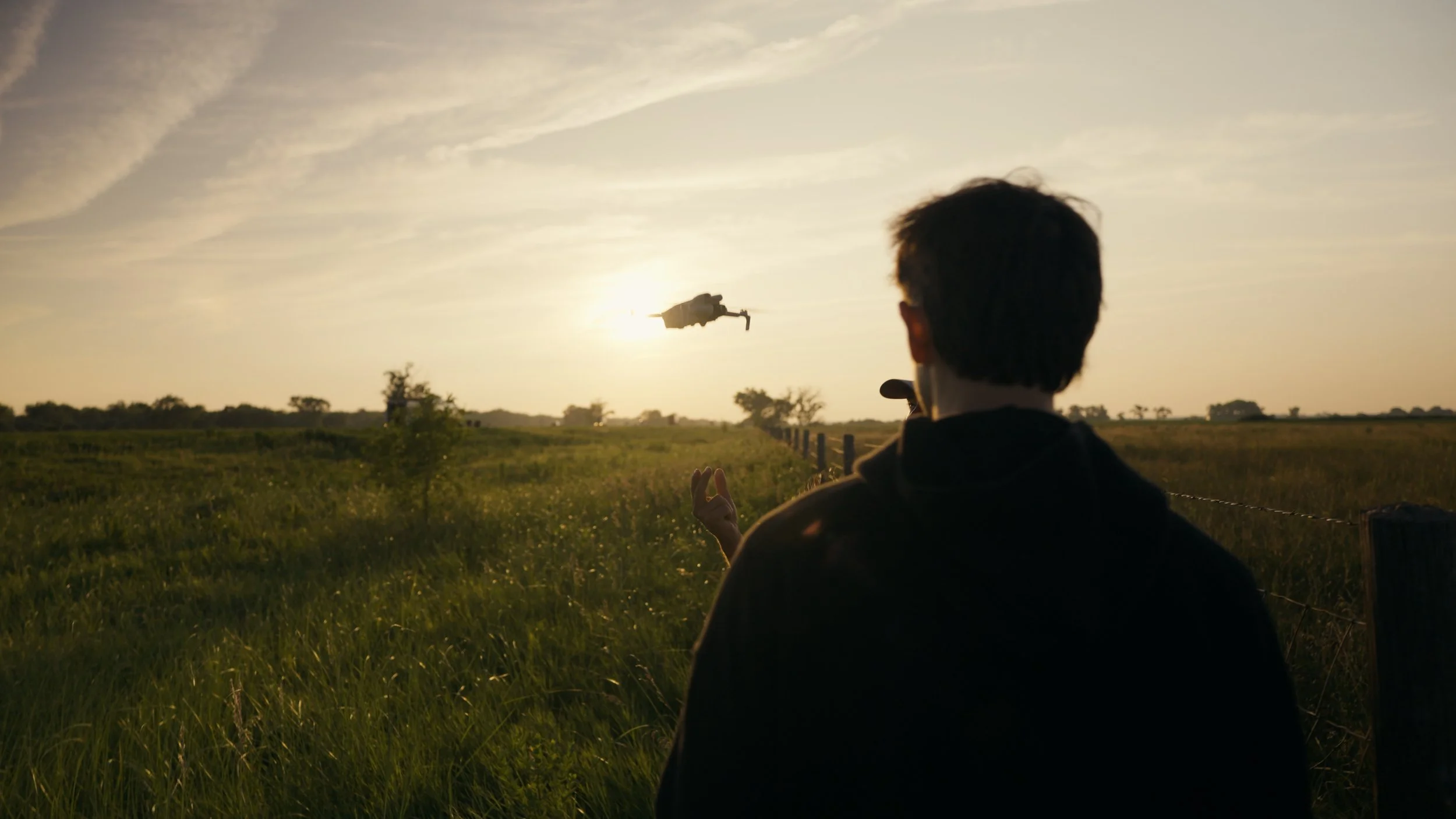 A person with dark hair and glasses is facing away, standing in a field at sunset, watching a drone fly in the sky.