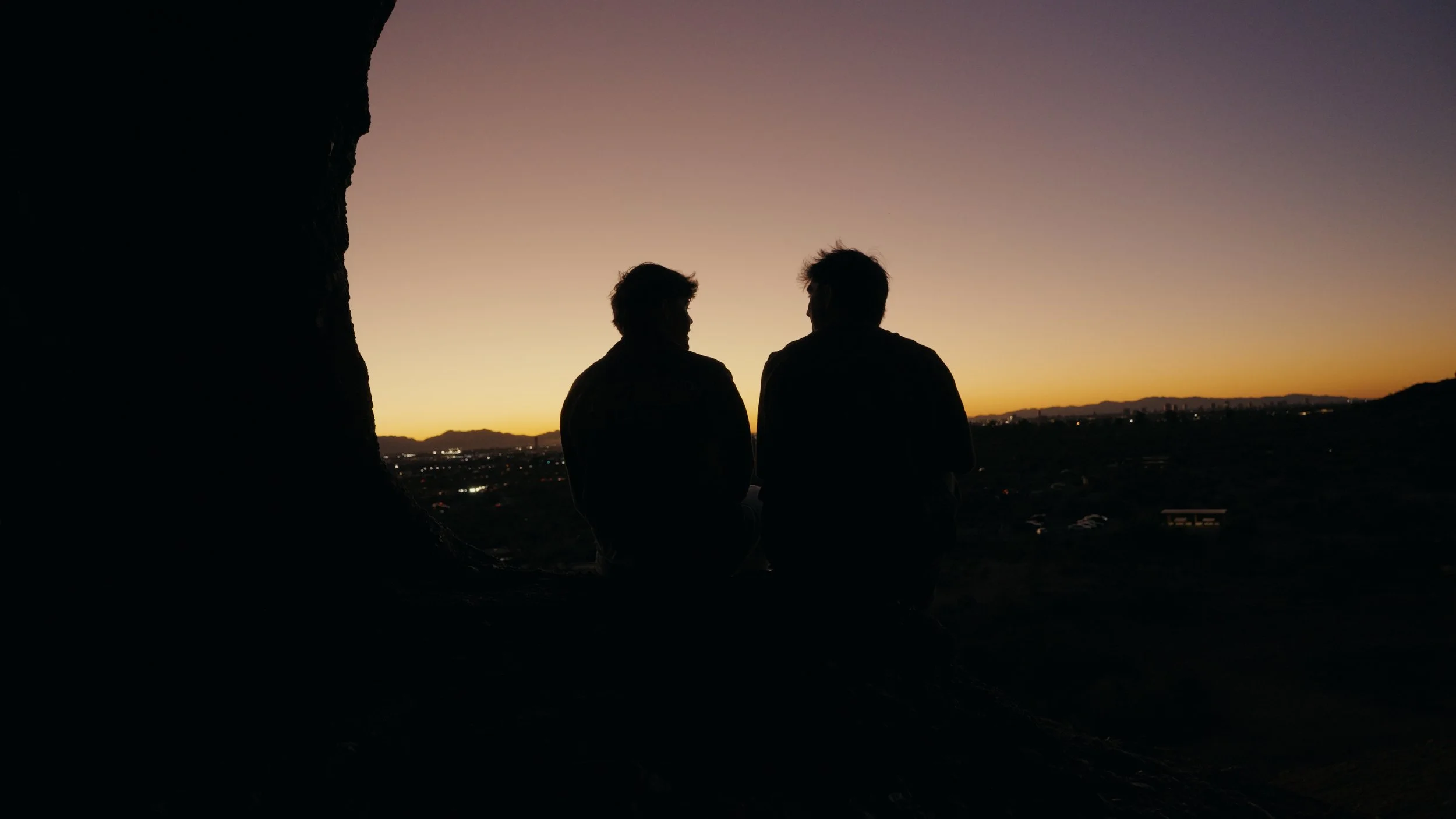 Silhouetted two people sitting on a ledge at sunset, with a dark foreground and a colorful sky in the background.