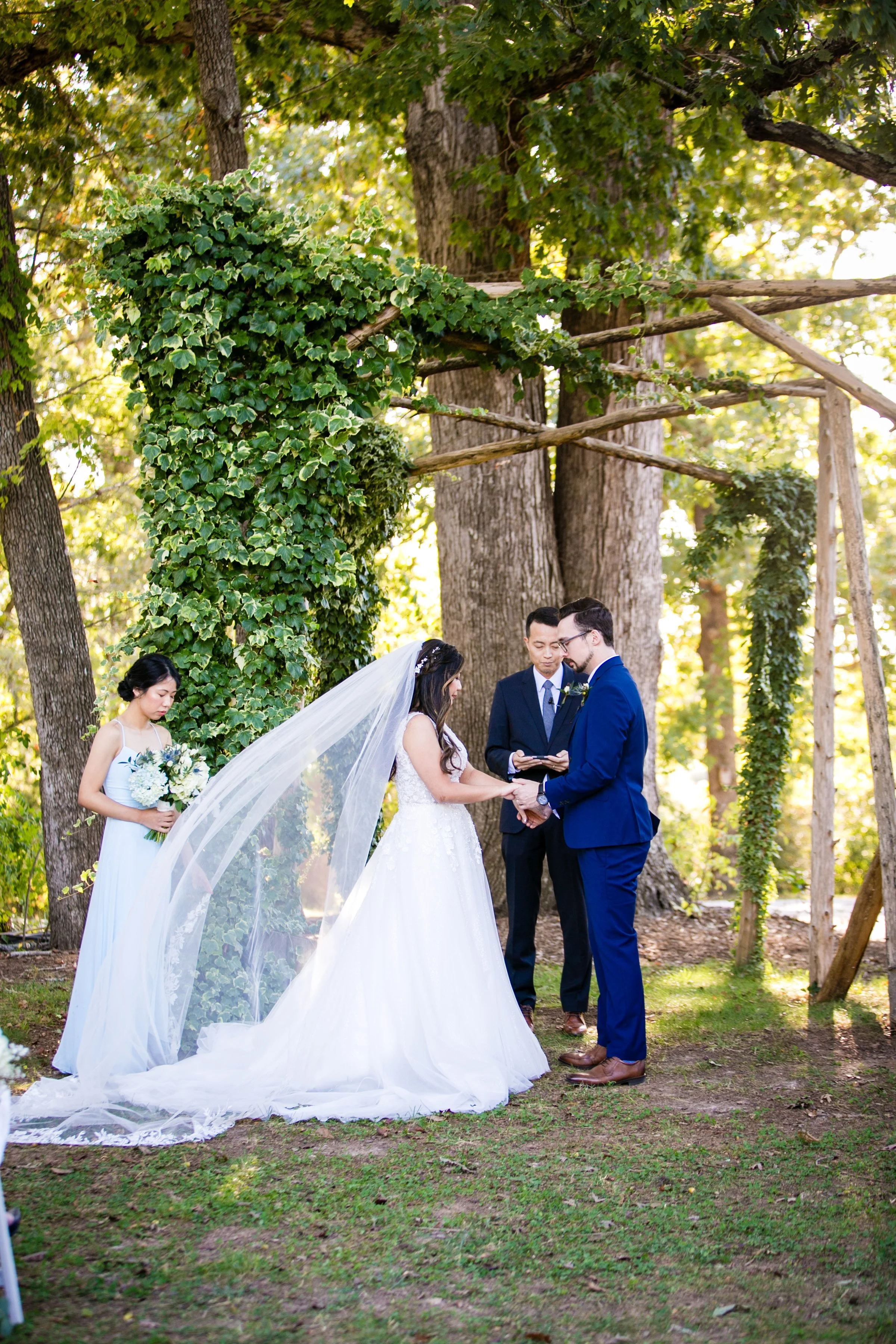 A wedding ceremony taking place outdoors in a forested area, with the bride and groom holding hands and exchanging vows, officiant, and bridesmaid with bouquet, under a wooden arch entwined with greenery.