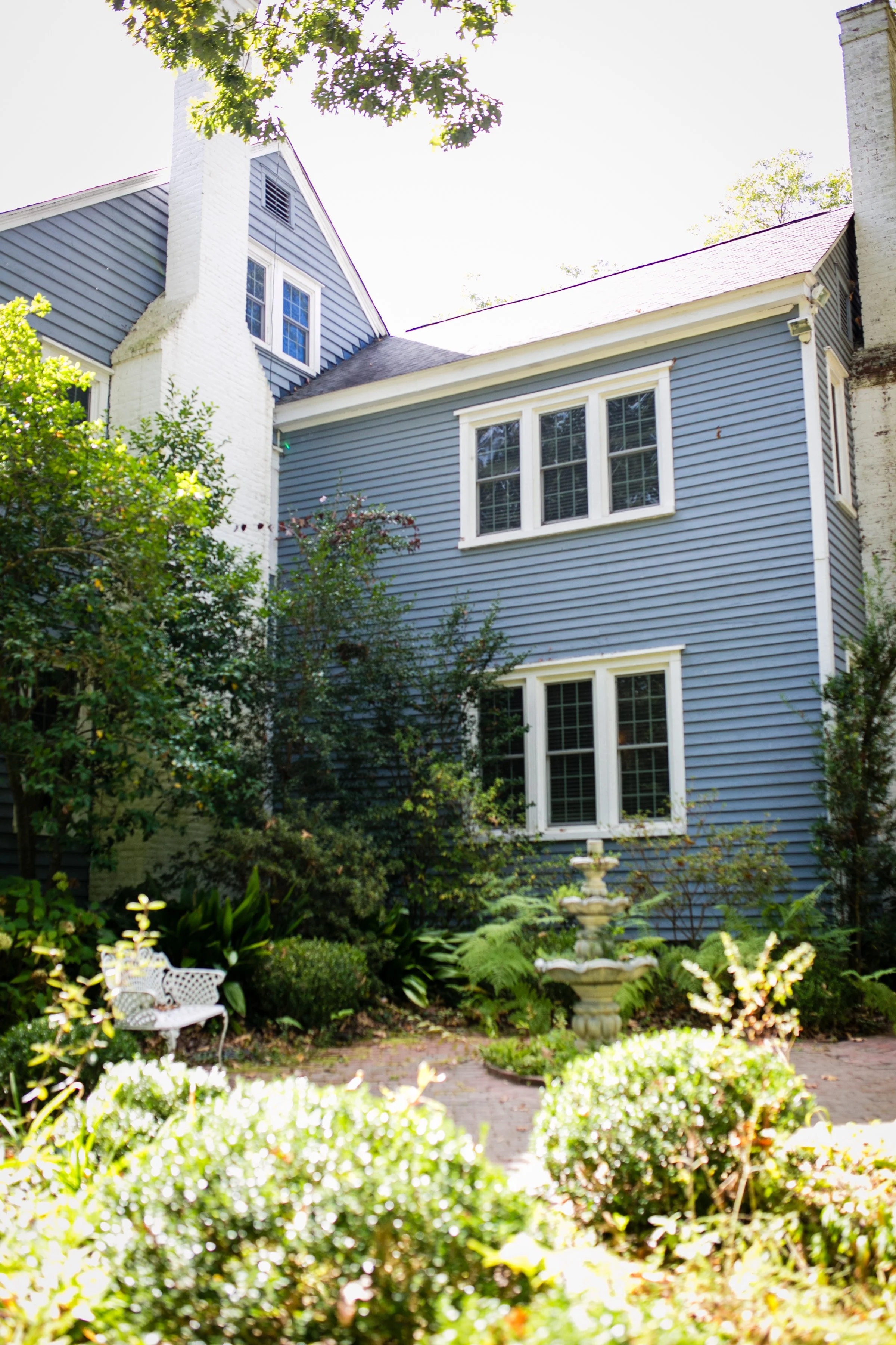 A blue house with white trim, three windows, and a garden in the foreground featuring lush green plants, bushes, and a stone fountain.