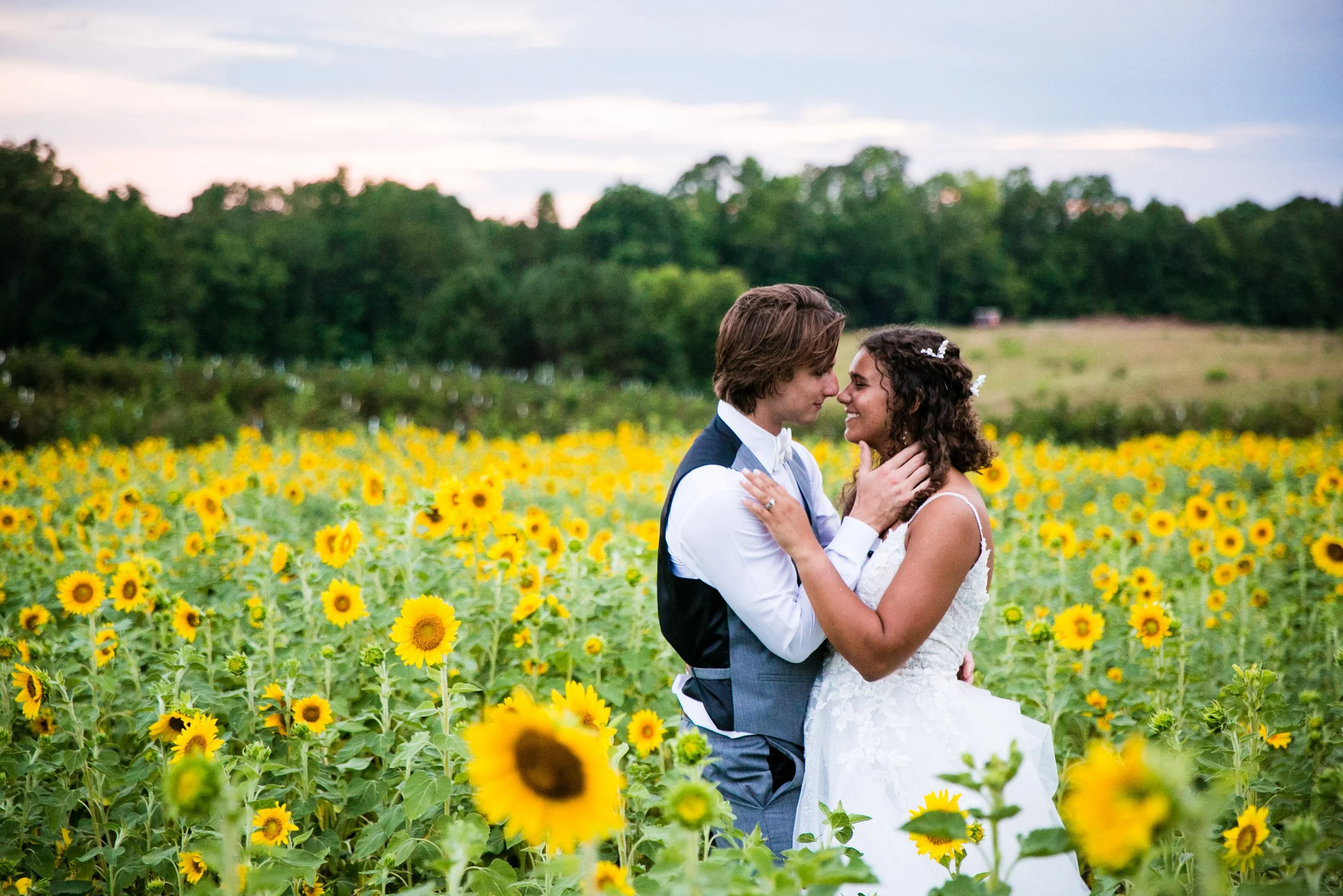 A couple dressed in wedding attire embracing in a sunflower field during sunset.