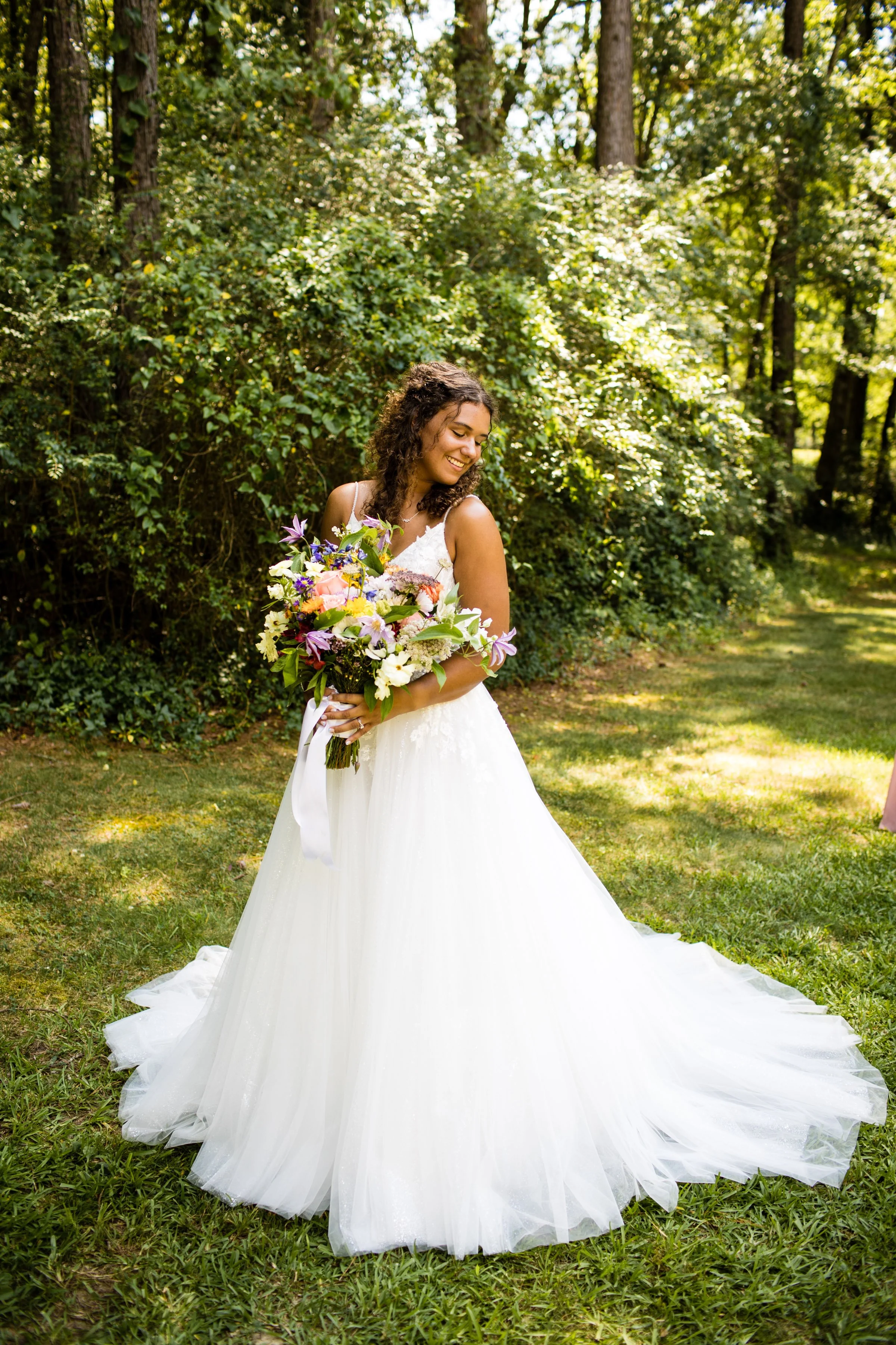 A bride in a white wedding gown holding a colorful bouquet of flowers, standing outdoors in a lush, green wooded area, smiling and looking down.