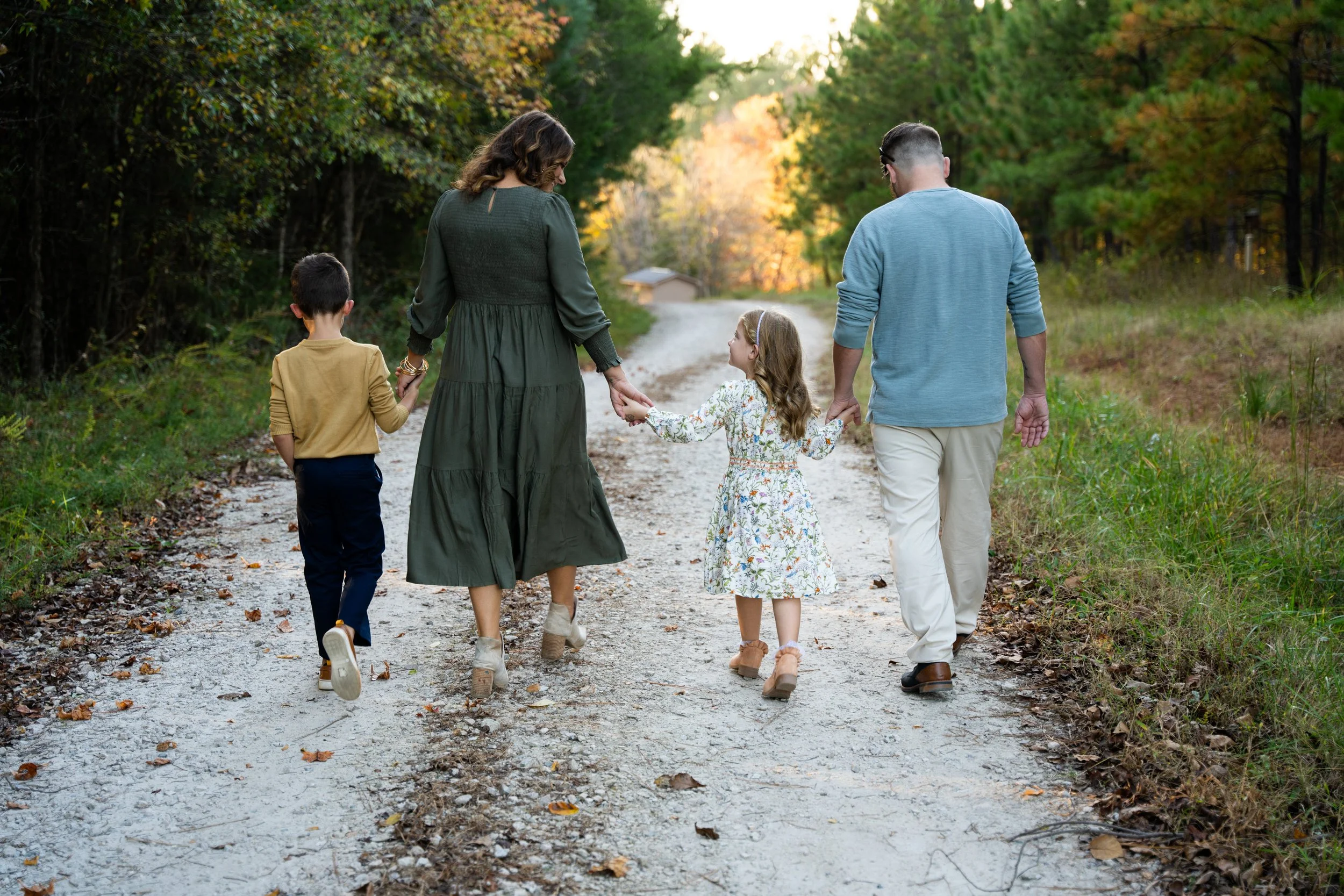 A family of five walking hand in hand on a dirt path in a forest during autumn. The family consists of a woman, a man, two young children, and a teenager, all dressed casually and enjoying a stroll through the fall foliage.
