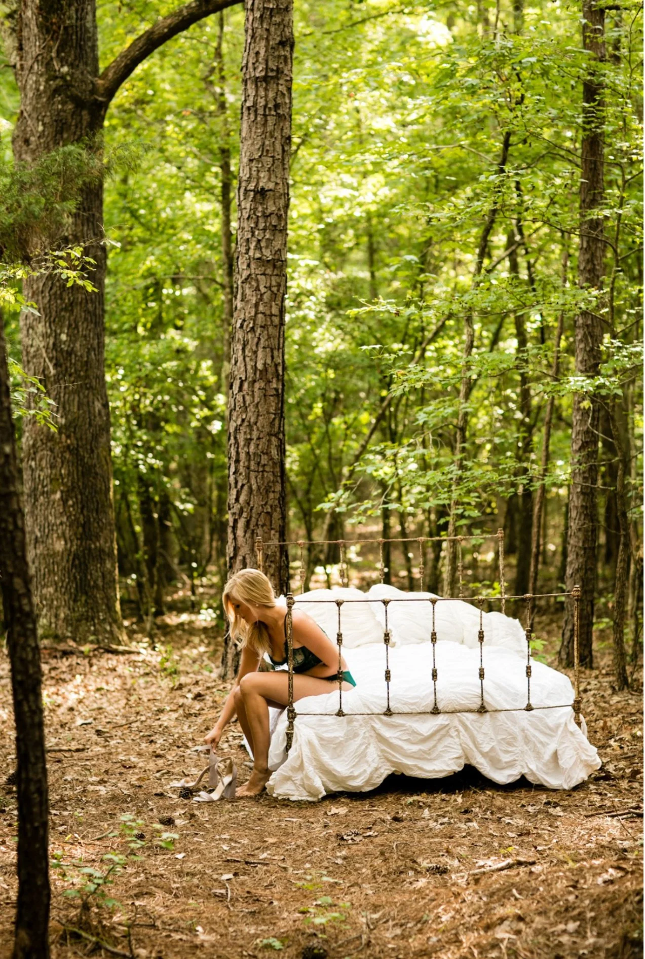 A woman sitting on a vintage metal-framed bed in a forest, putting on high-heeled shoes, surrounded by tall trees and green foliage.