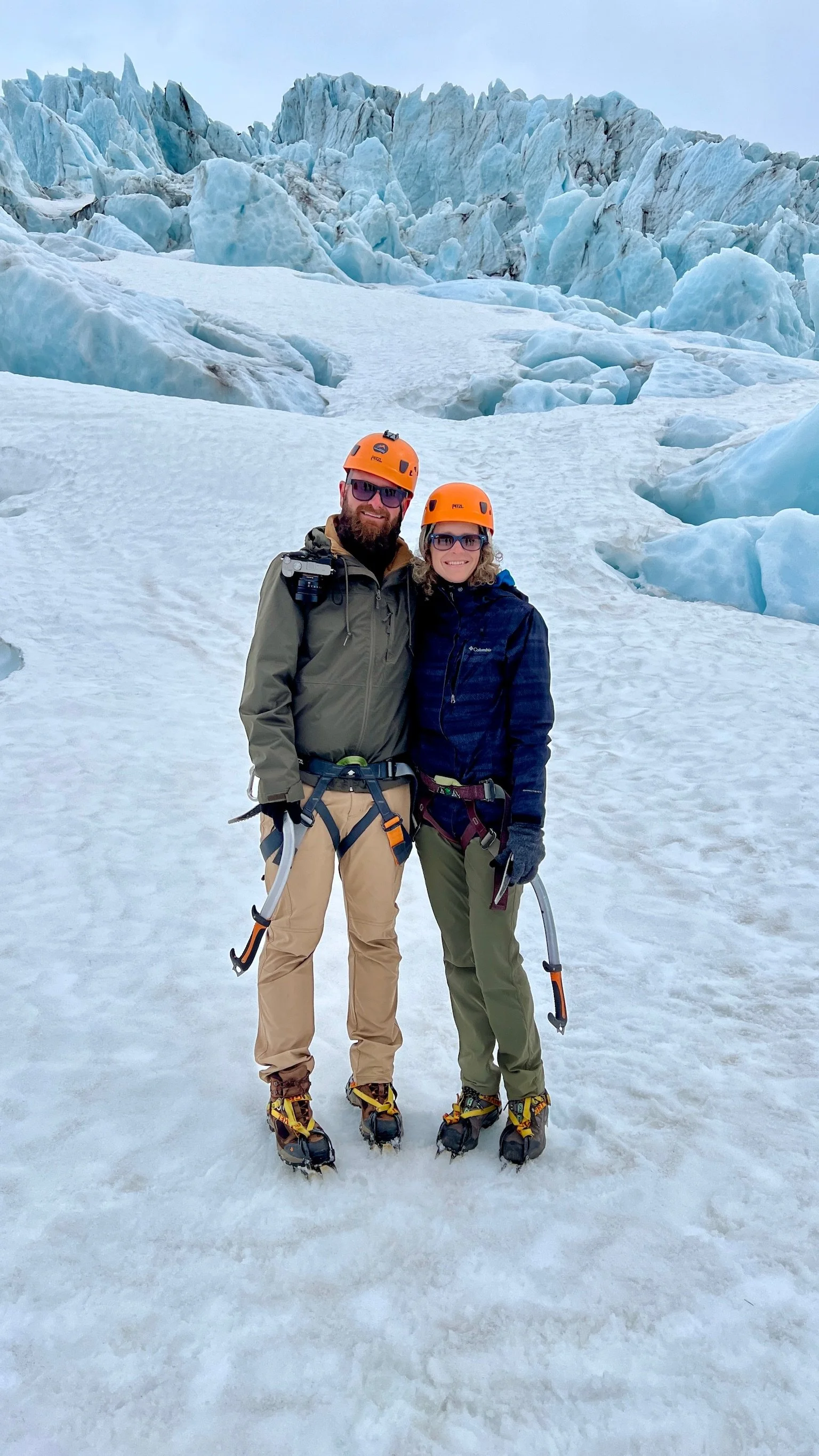 Two people wearing helmets and ice axes stand on snow-covered ice in front of a glacier, dressed in outdoor gear and ice climbing equipment.