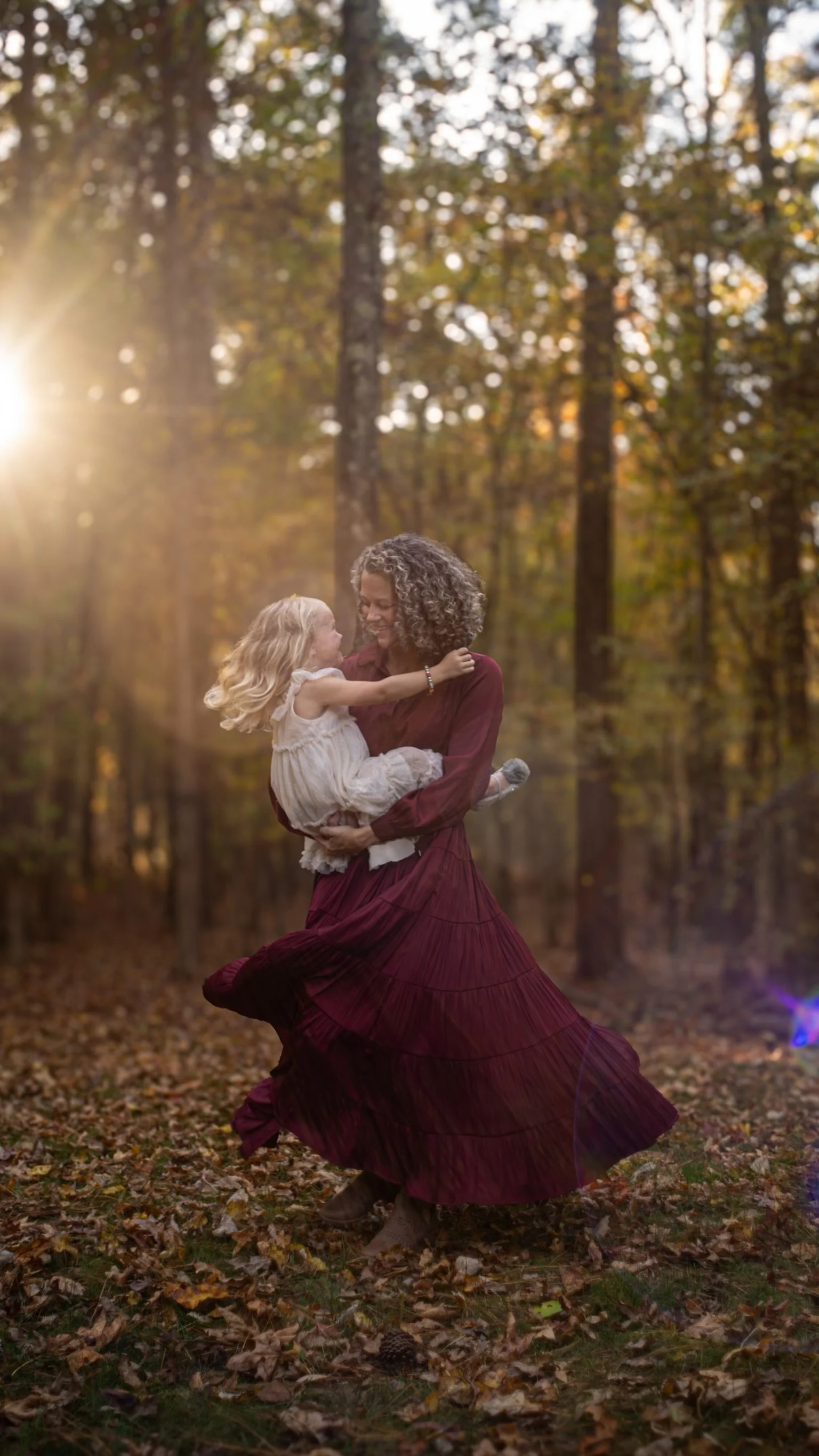 A woman wearing a maroon dress joyfully spins while holding a young girl in a white dress in a forest setting during autumn, with sunlight filtering through trees.