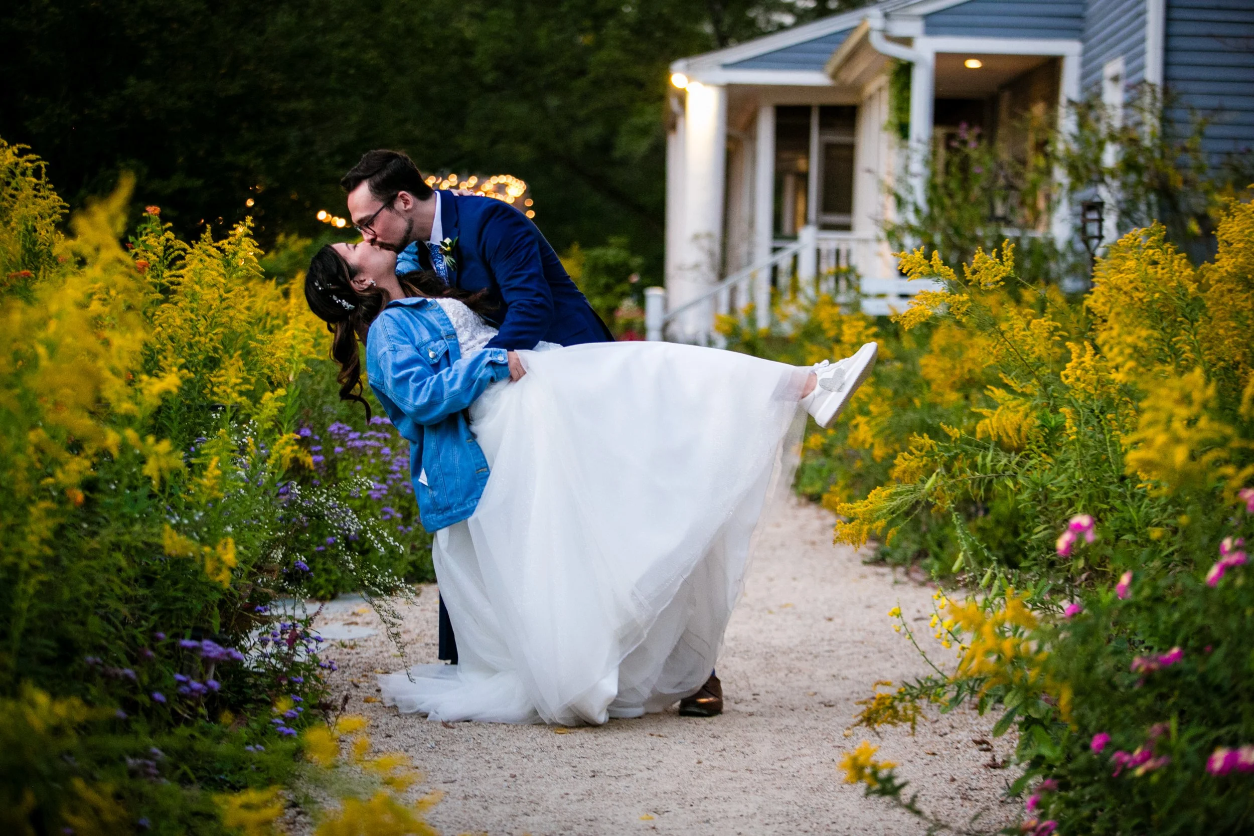 A newlywed couple in wedding attire sharing a kiss in a garden aisle surrounded by yellow and purple flowers, with a house in the background.
