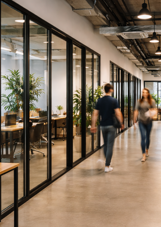 Modern coworking office corridor with glass-walled meeting rooms, indoor plants, and two people walking.