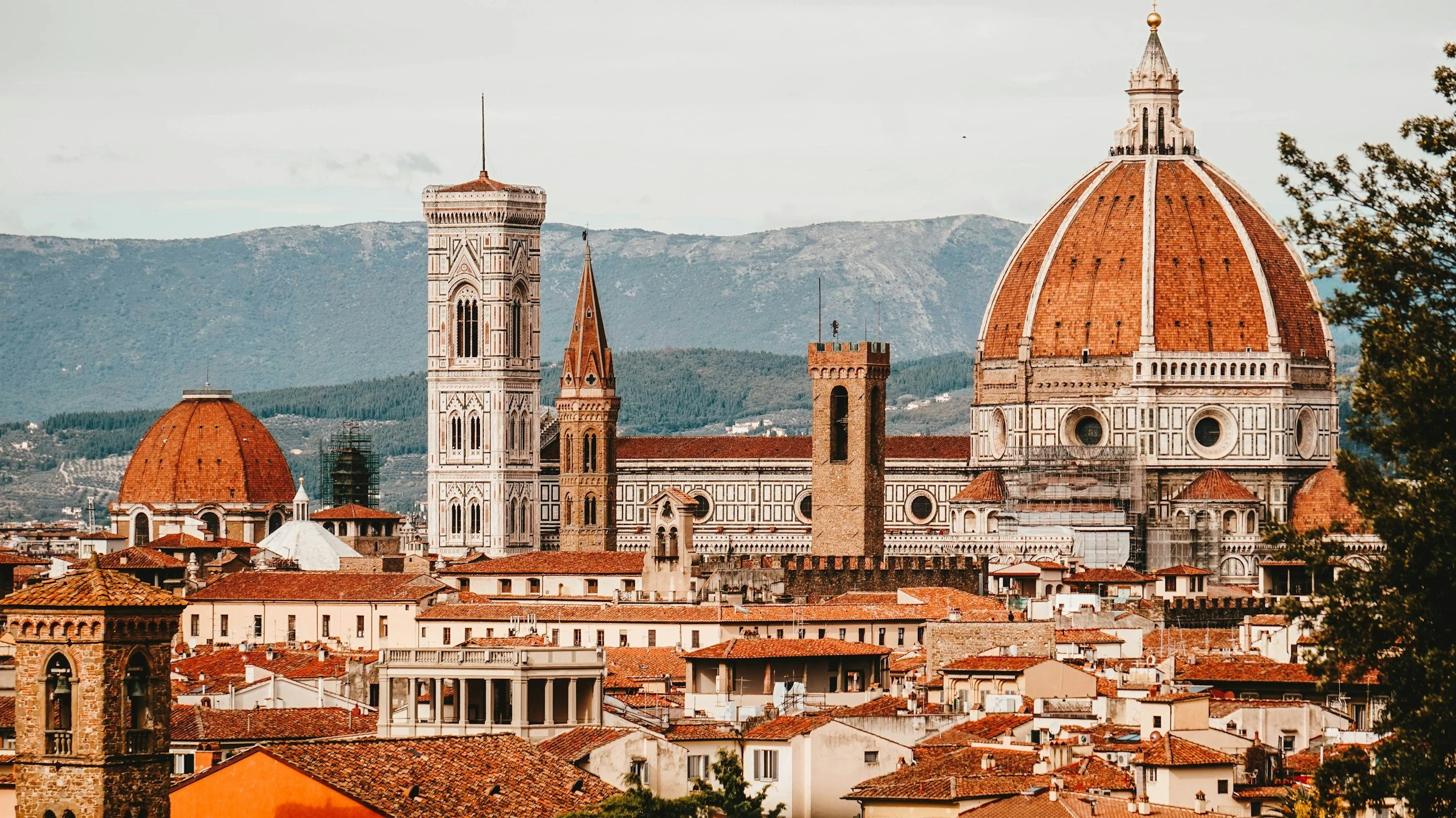 Panoramic view of Florence, Italy with the Florence Cathedral (Duomo) and other historic buildings and rooftops in the foreground, and mountains in the background.