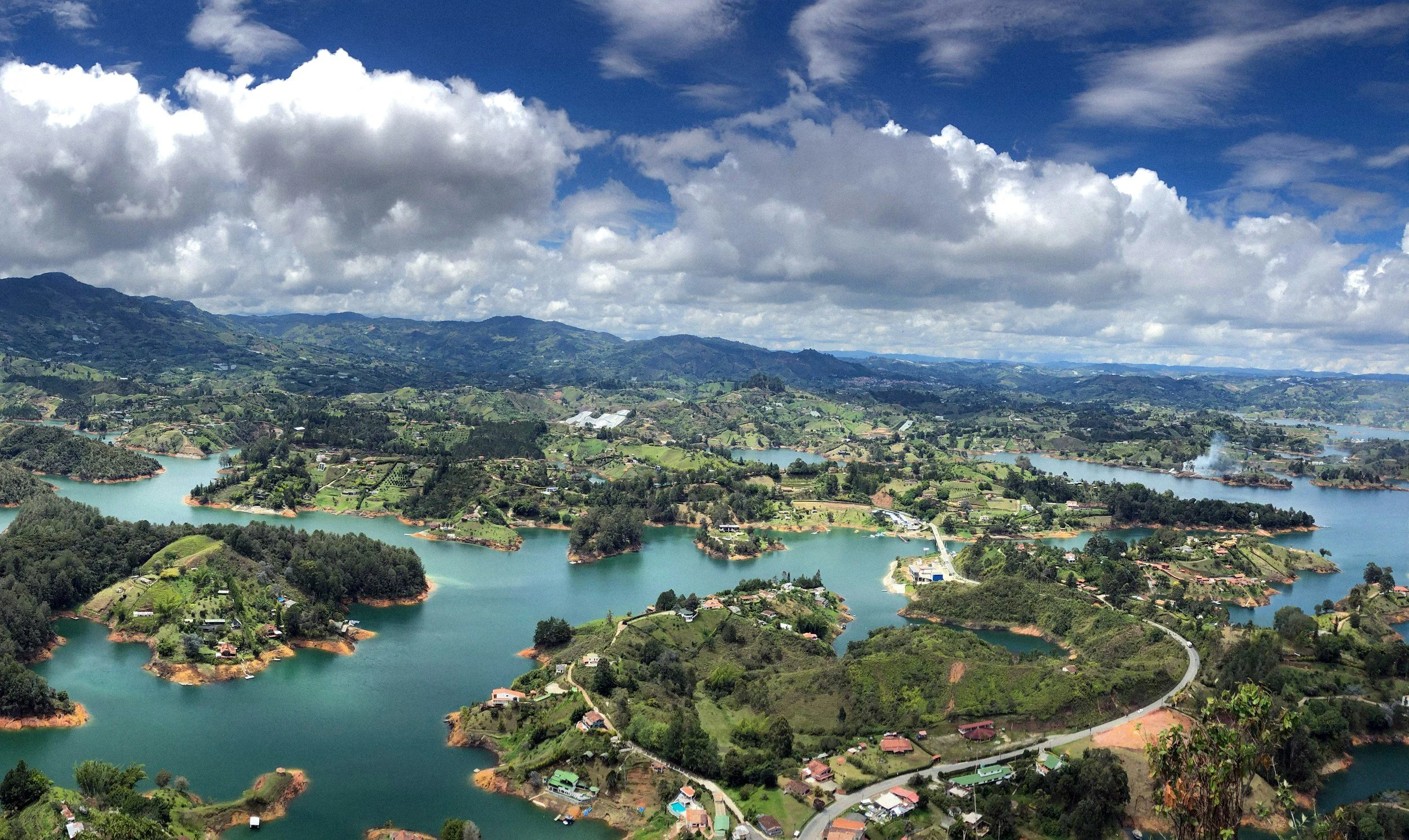 Aerial view of a landscape with multiple lakes, green hills, scattered houses, and winding roads under a partly cloudy sky.