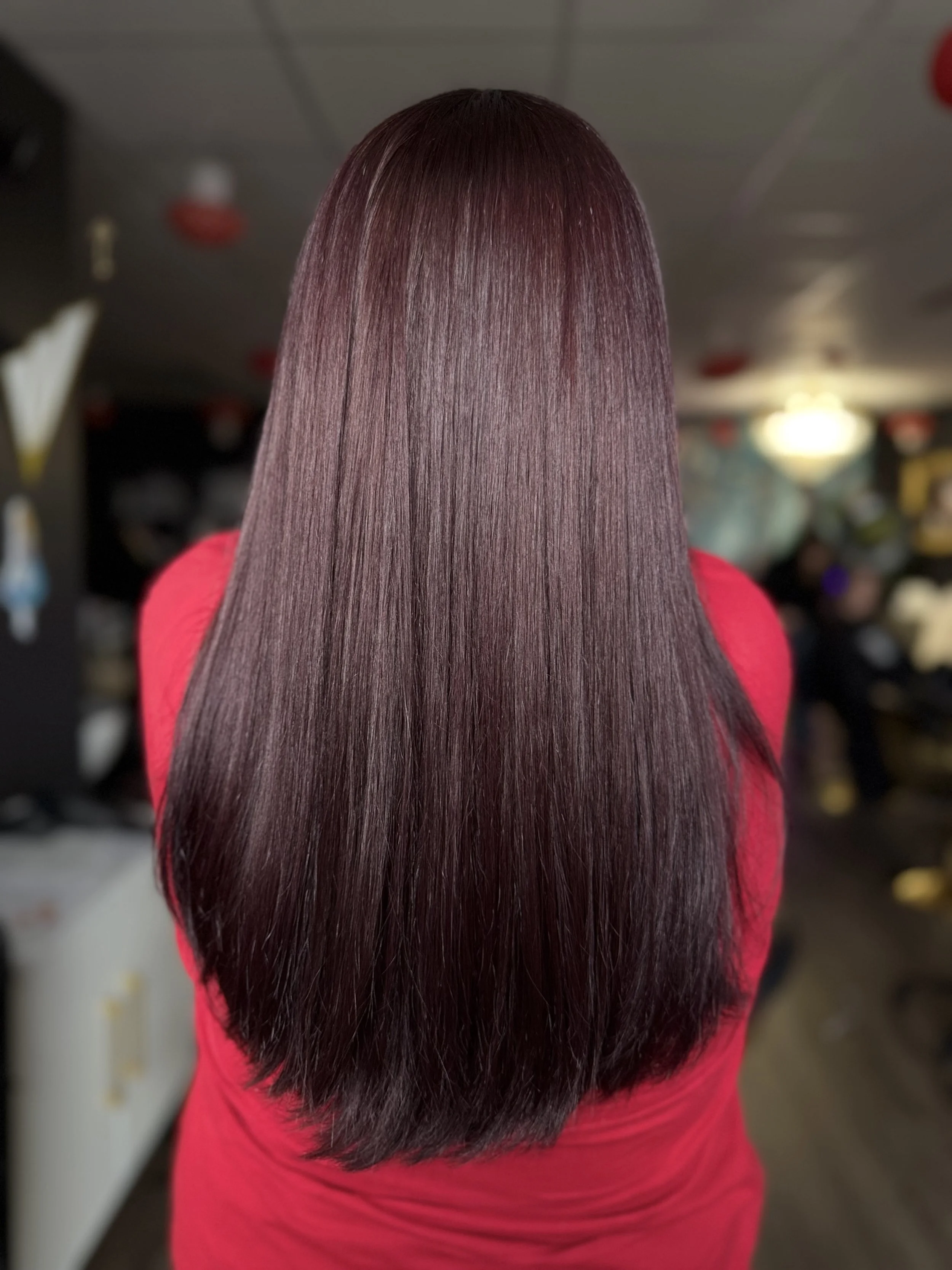 Back of a woman with long, straight, shiny dark red hair, wearing a red shirt, in a salon or similar setting.