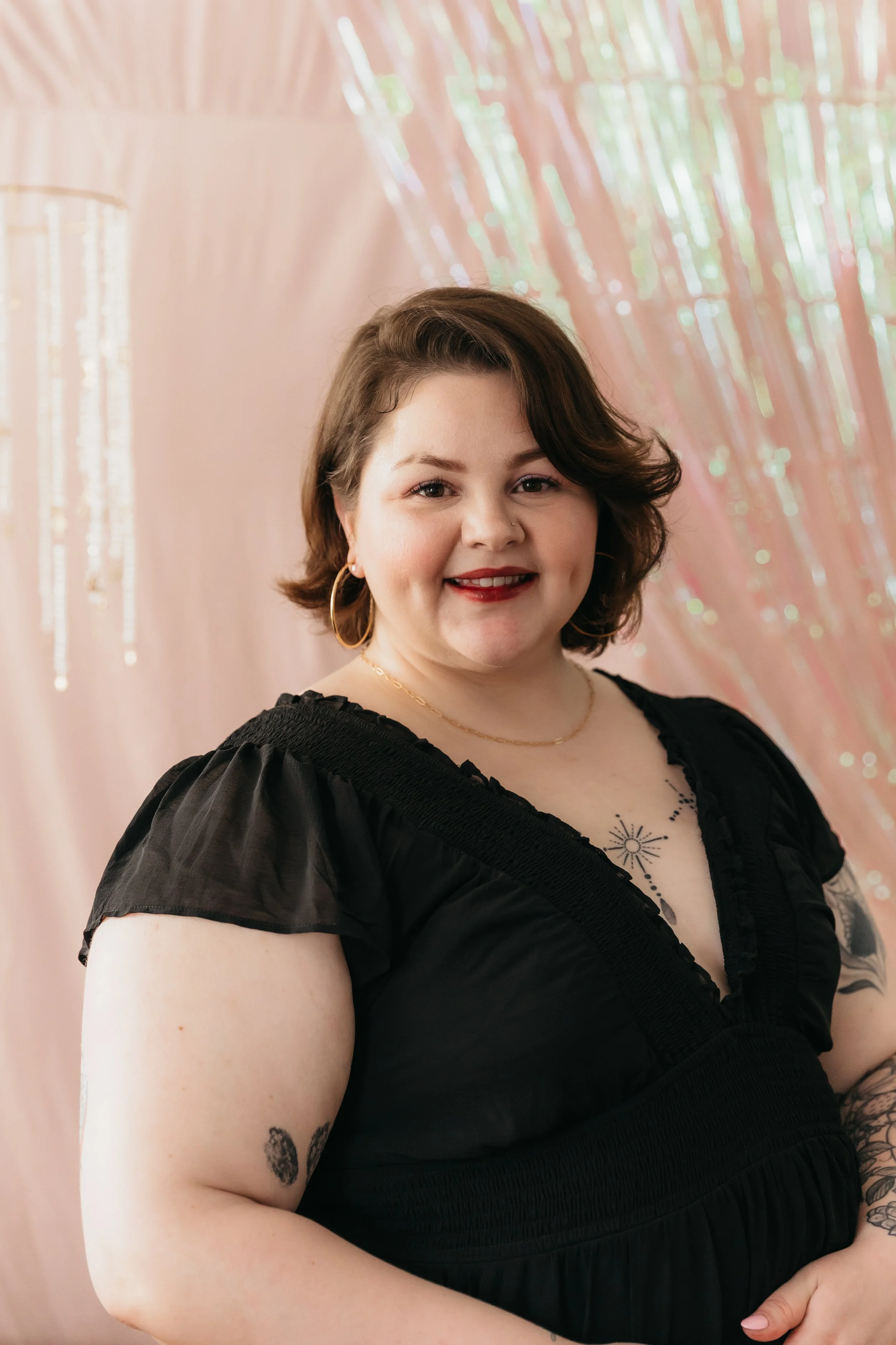 A woman with short brown hair, wearing a black dress with ruffled sleeves, gold hoop earrings, and a gold chain necklace, smiling in front of a pink and iridescent backdrop.