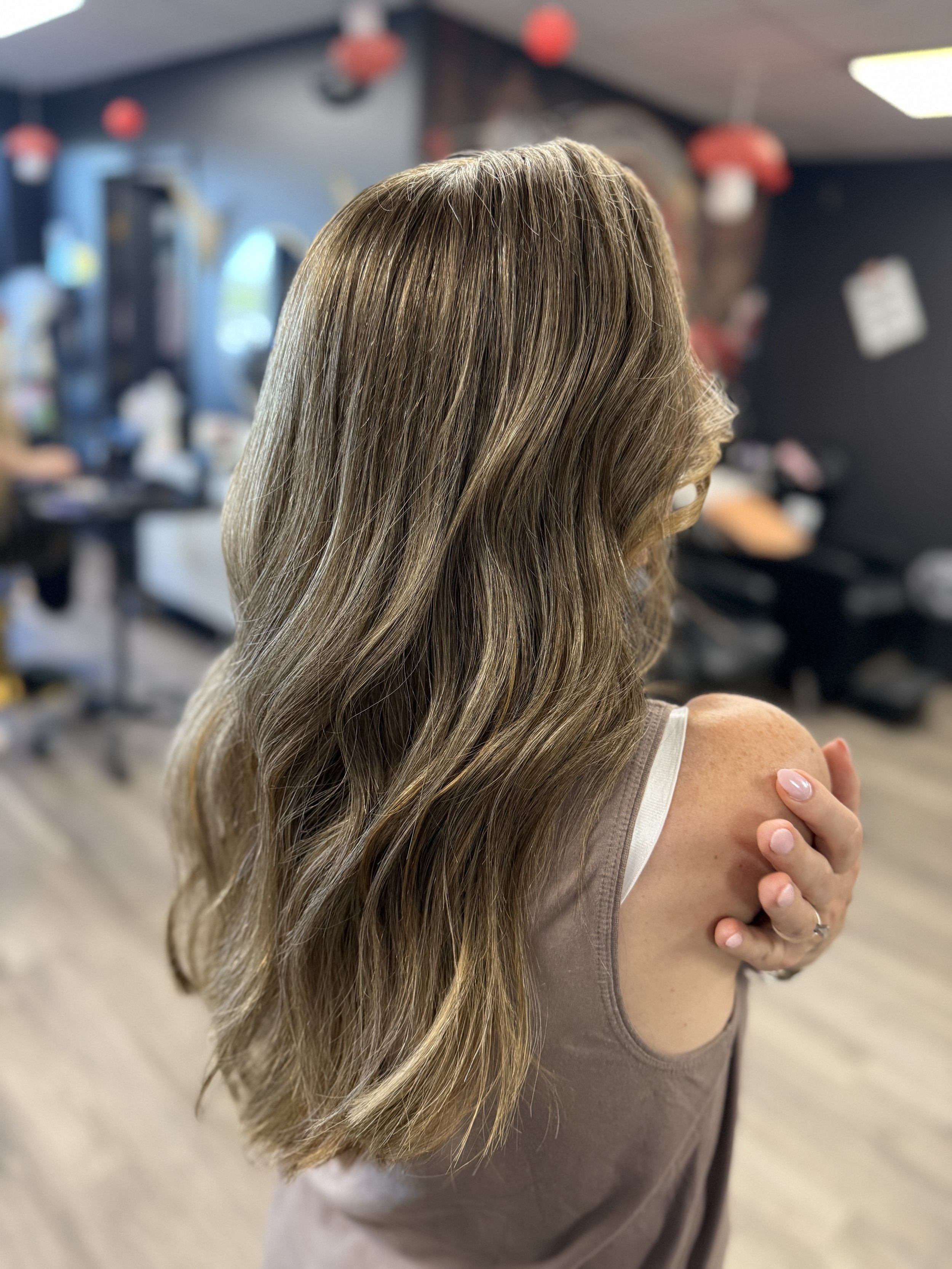 A woman with wavy, light brown hair standing in a salon, touching her shoulder, with a blurred salon background.