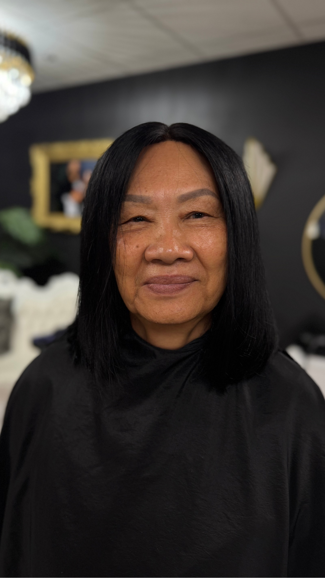 A middle-aged woman with straight black hair, smiling softly, wearing a black top, standing indoors with a dark wall, framed art, and decorative mirror in the background.
