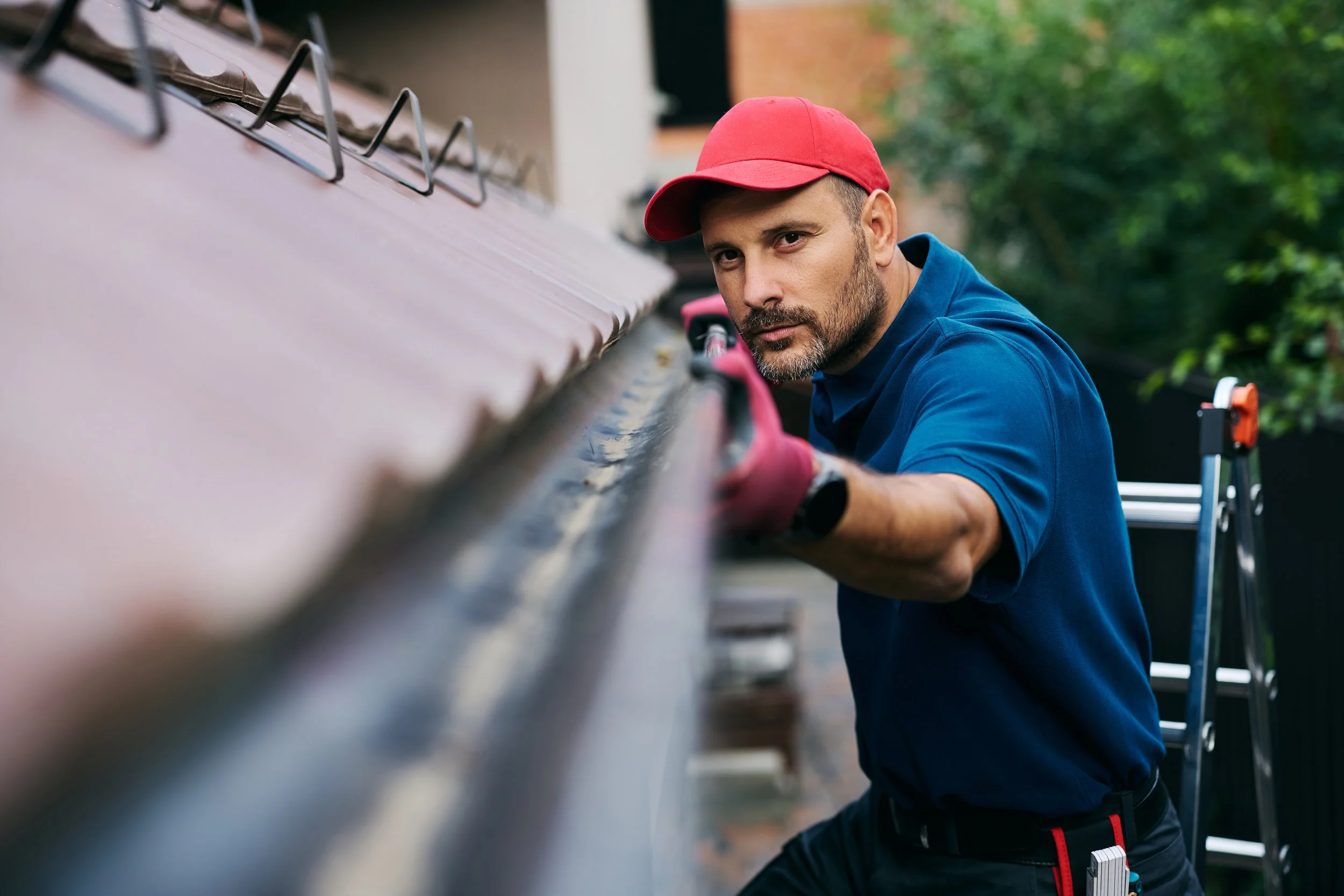 A man in a red cap and blue shirt fixing the roof of a house, standing on a ladder outdoors.