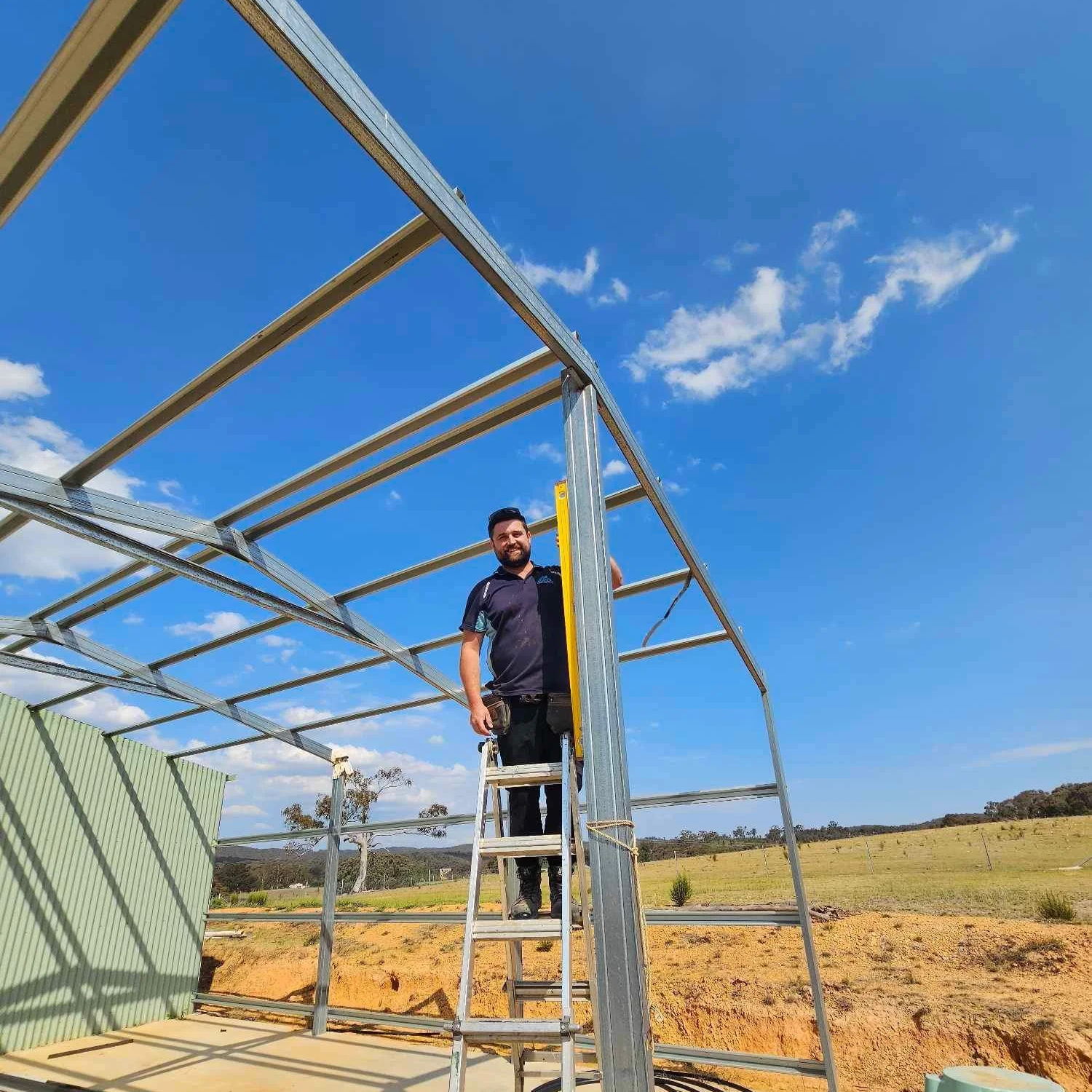 A man standing on a ladder working on a metal frame structure outdoors under a blue sky with scattered clouds.