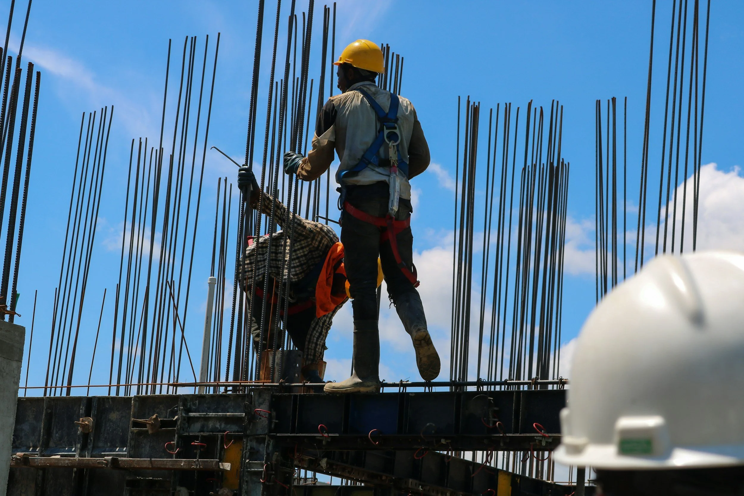 Construction workers on a building site, wearing safety gear and working among steel rebar, with a blue sky in the background.