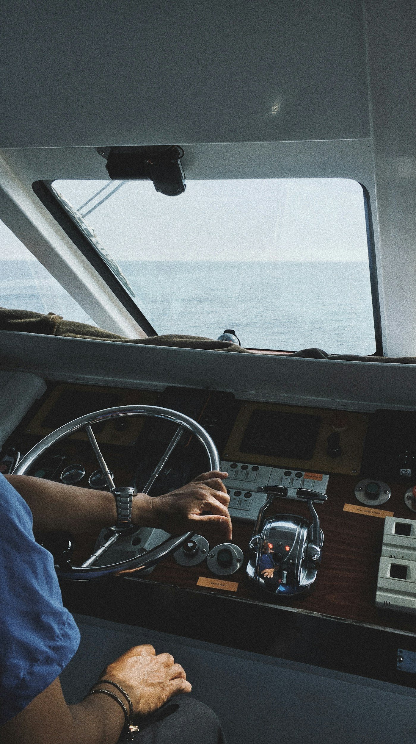 Inside the control room of a boat or ship, with a person operating the steering wheel, controlling navigation instruments, and looking out at the ocean through a large window.