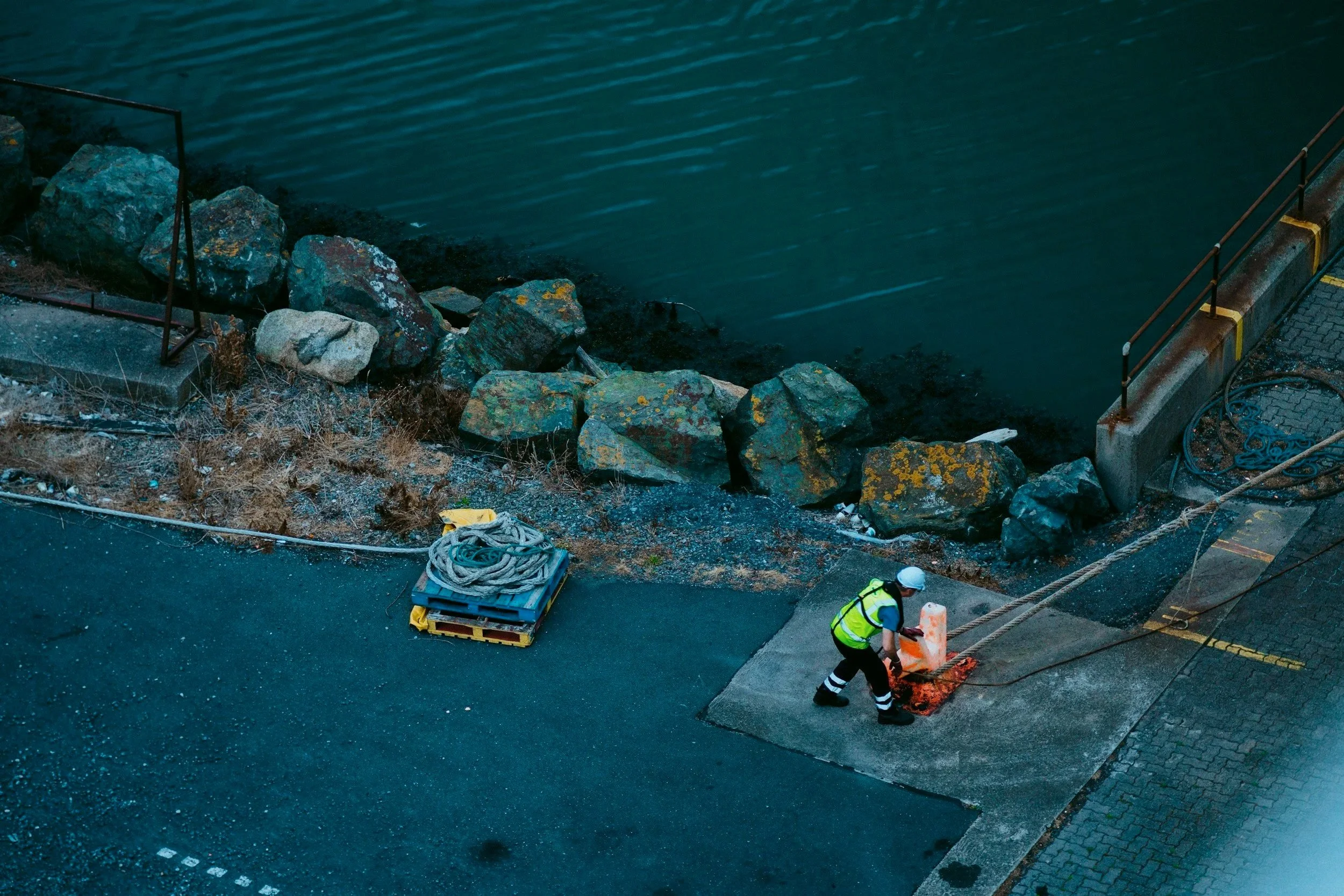 Worker in safety vest and helmet working near a waterway at night, with large rocks, a dock, and construction equipment nearby.