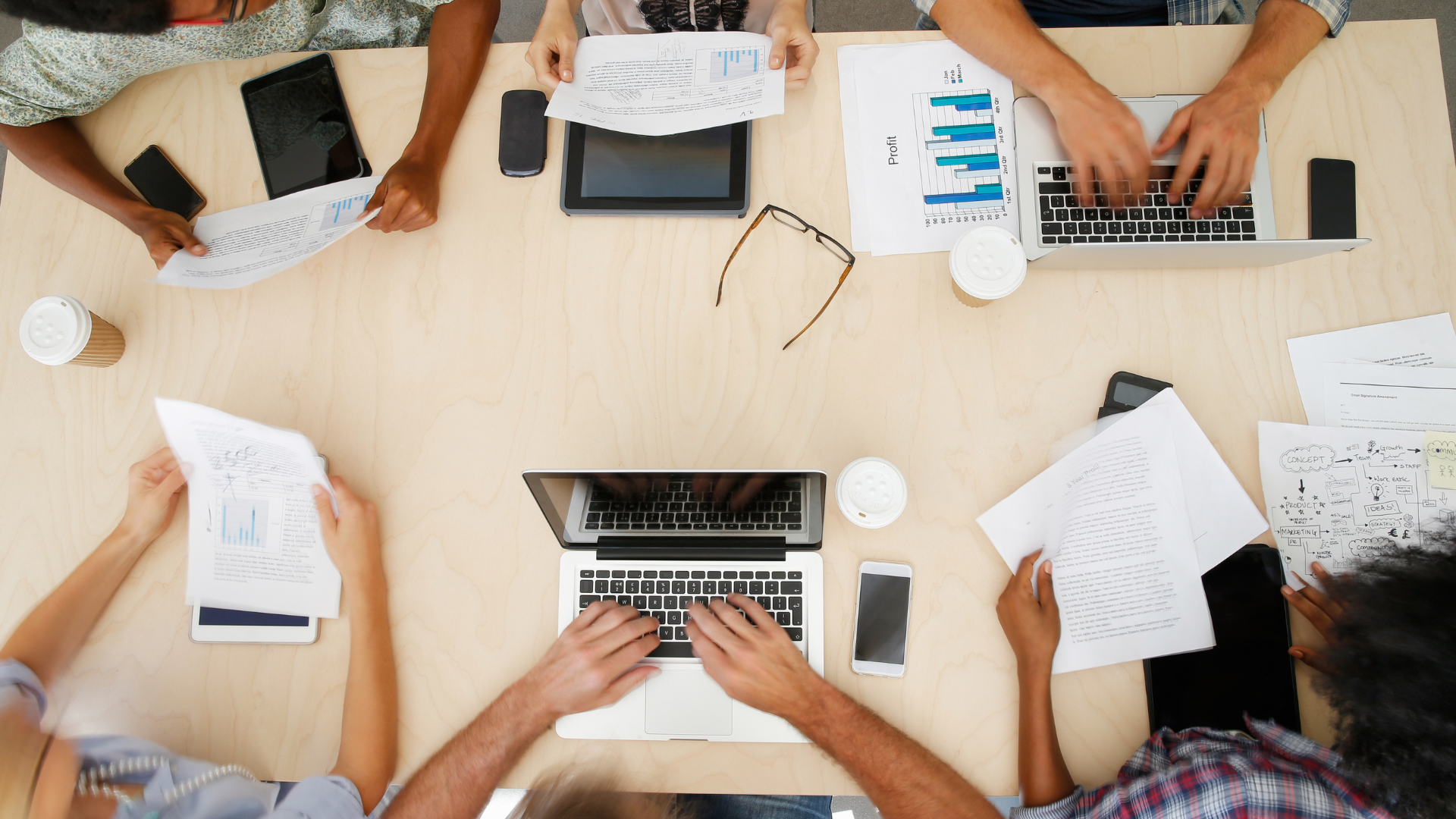 Top-down view of five people working at a light-colored wooden table with laptops, smartphones, printed documents with graphs, a tablet, a pair of glasses, and coffee cups.