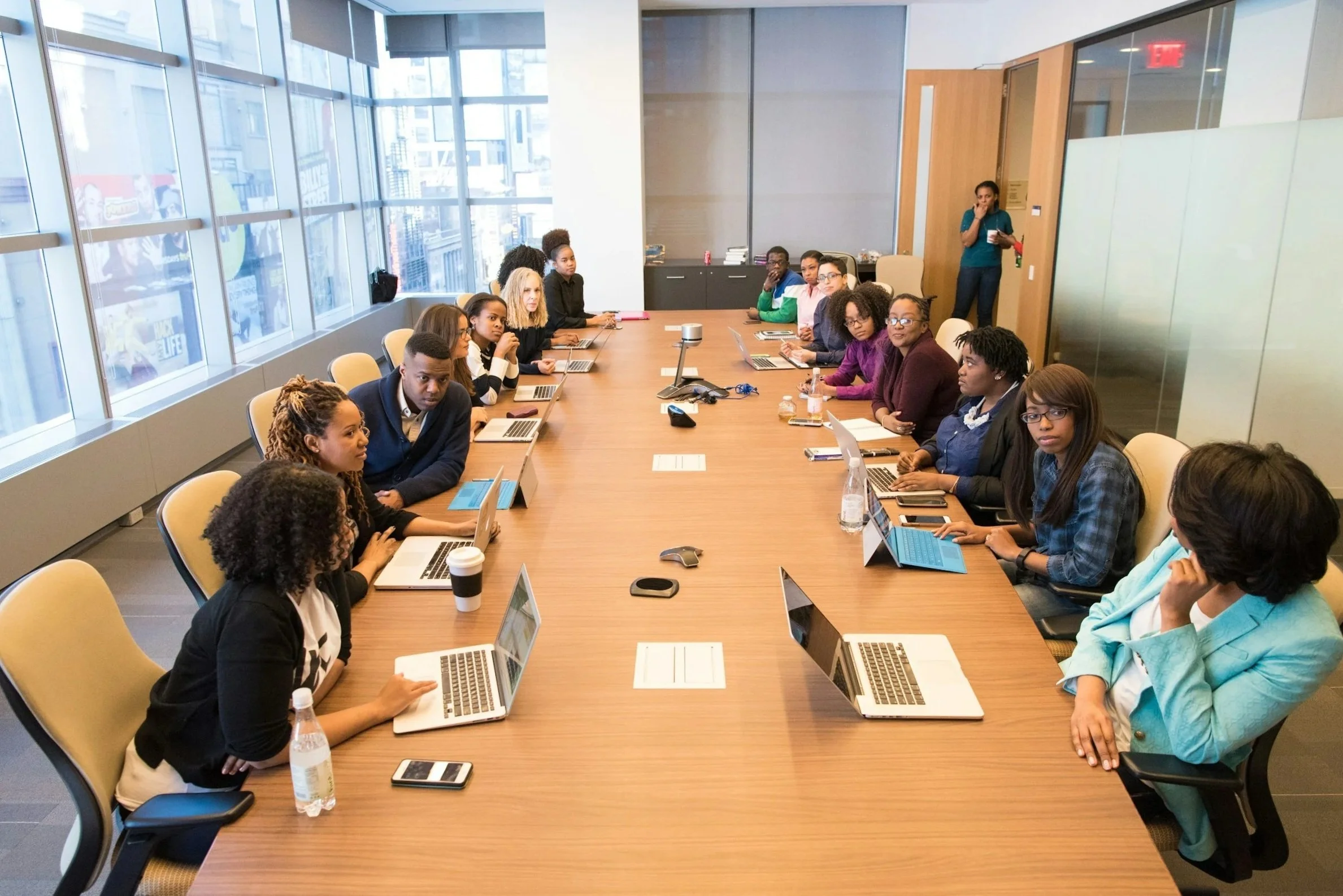 A diverse group of professionals in a modern classroom, some working on laptops, gathered around a large conference table during a meeting.