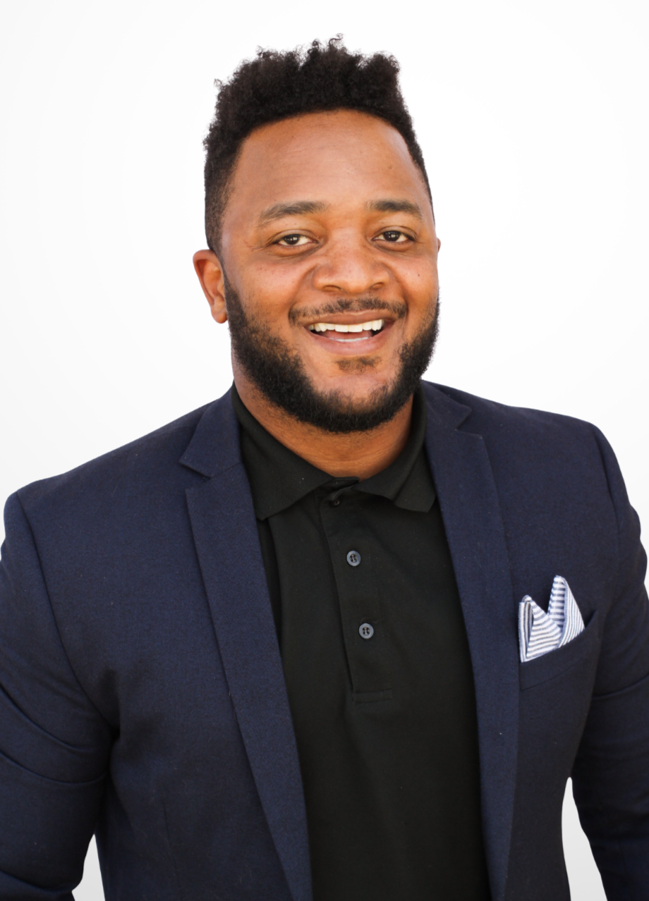 Smiling man with dark hair and beard wearing a navy blazer and black shirt, with a patterned pocket square, standing against a white background.