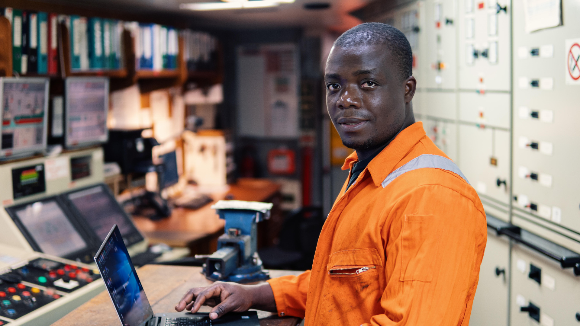 A man in an orange safety uniform working in an electrical control room with multiple control panels and computer screens.