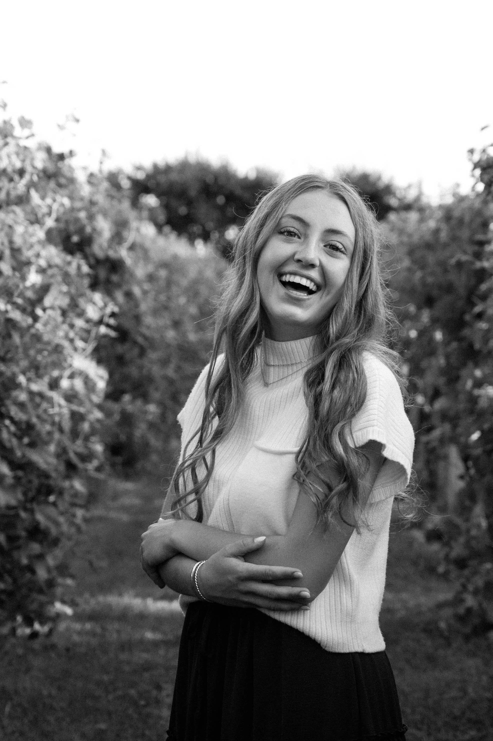 A young woman smiling outdoors, standing amidst foliage, wearing a short-sleeve sweater and a skirt.