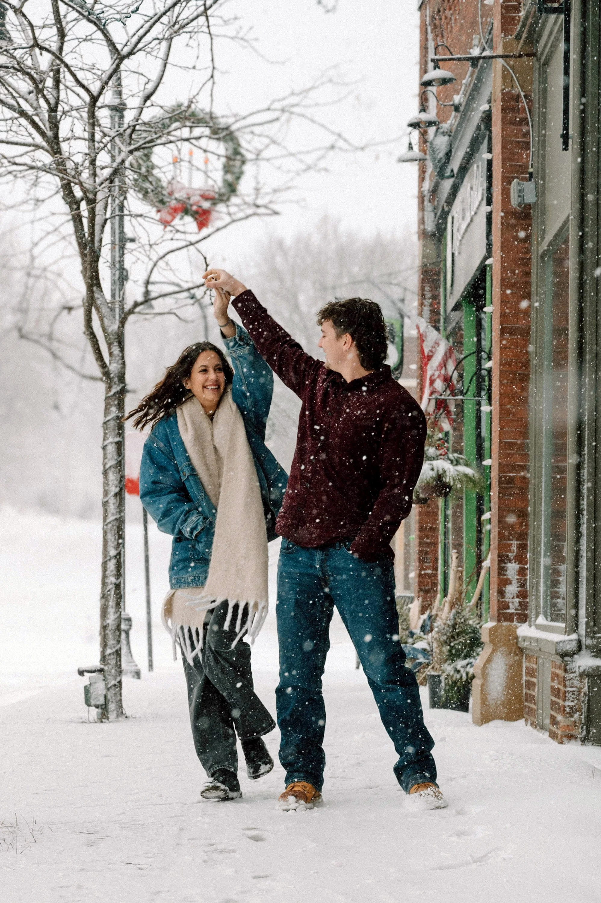 A young couple dancing in the snow on a winter day, with a basketball hoop and decorated storefront in the background.