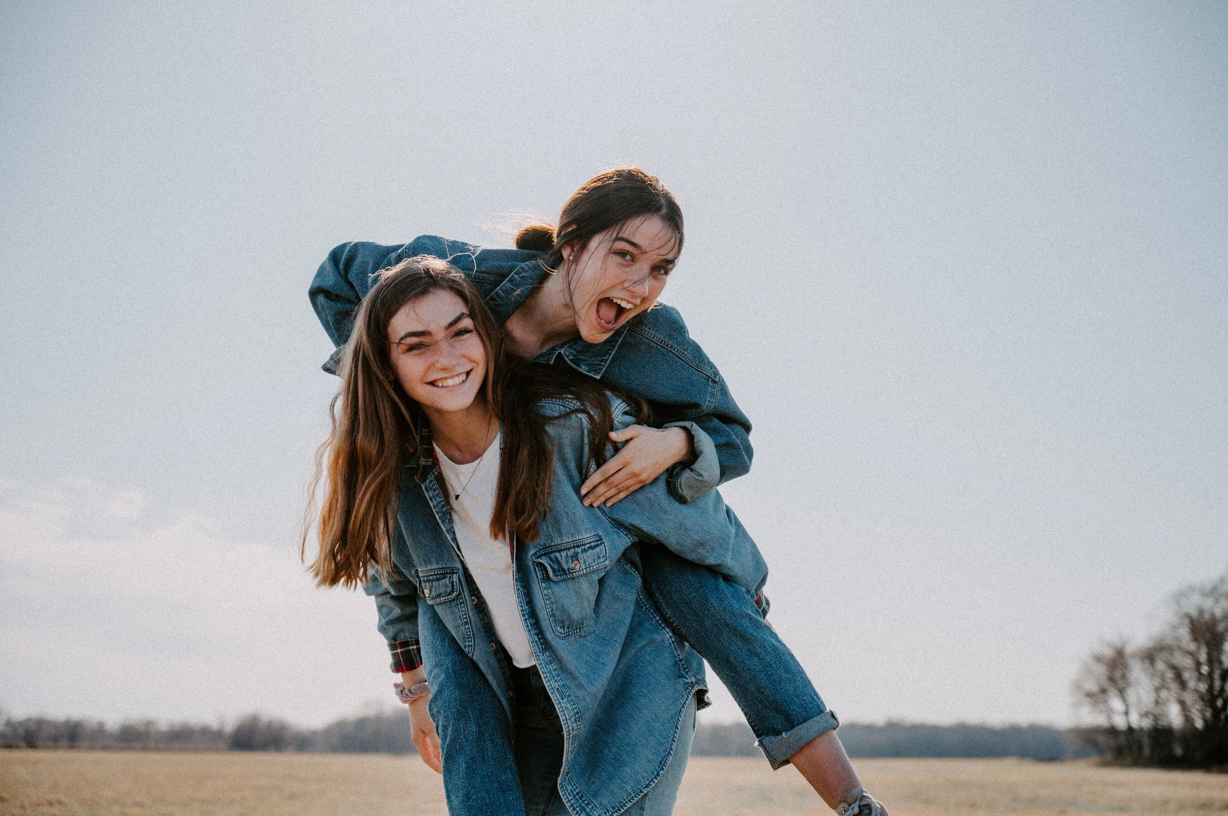 Two young women in denim jackets and jeans enjoying outdoors on a clear day. One is giving the other a piggyback ride, and they are both smiling and laughing.