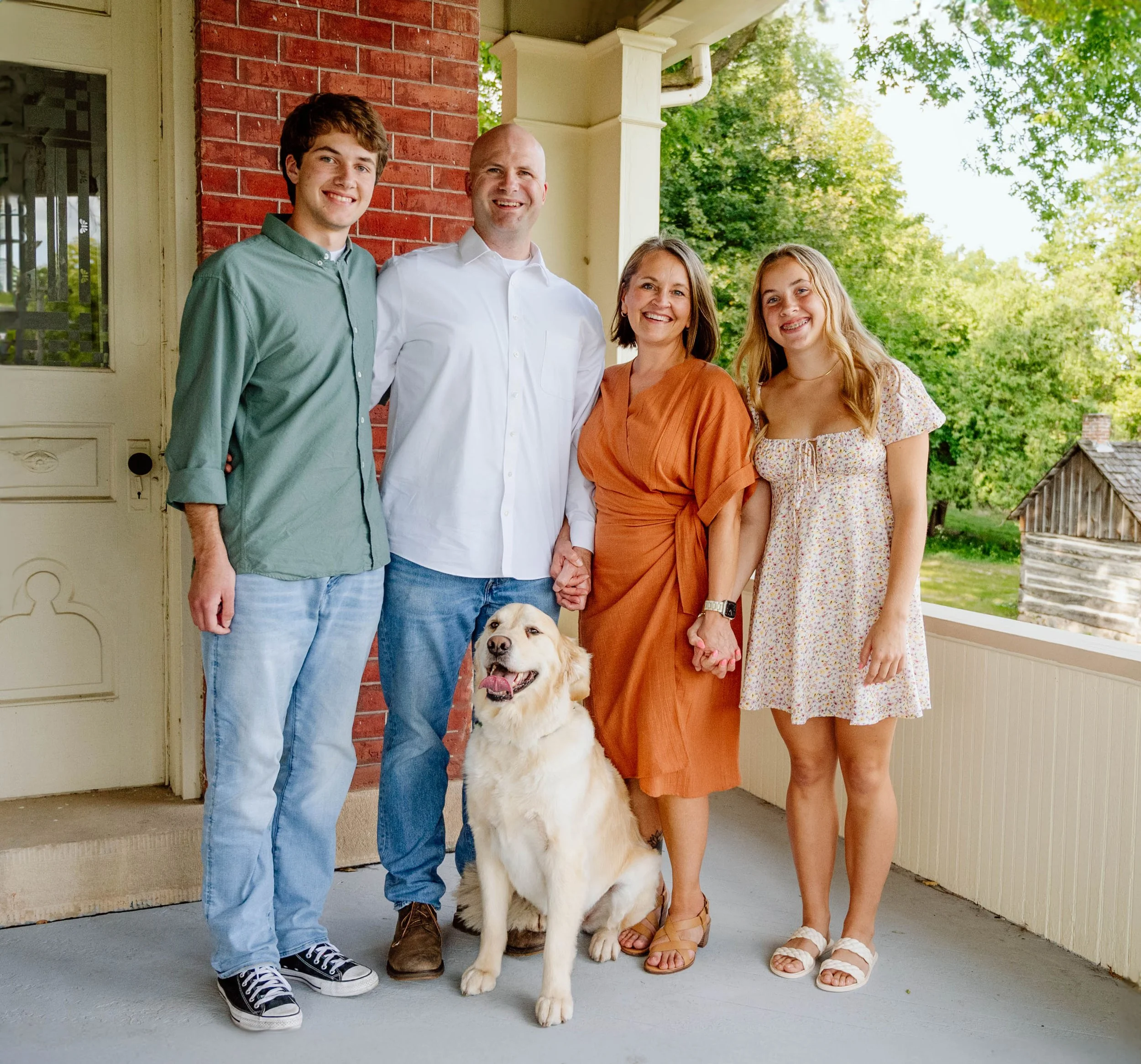A family of four and a dog standing on a porch. The family includes two young women, a middle-aged woman, and a middle-aged man. They are smiling and holding hands, with a dog sitting in front of them. The setting is outdoors with trees and a house v